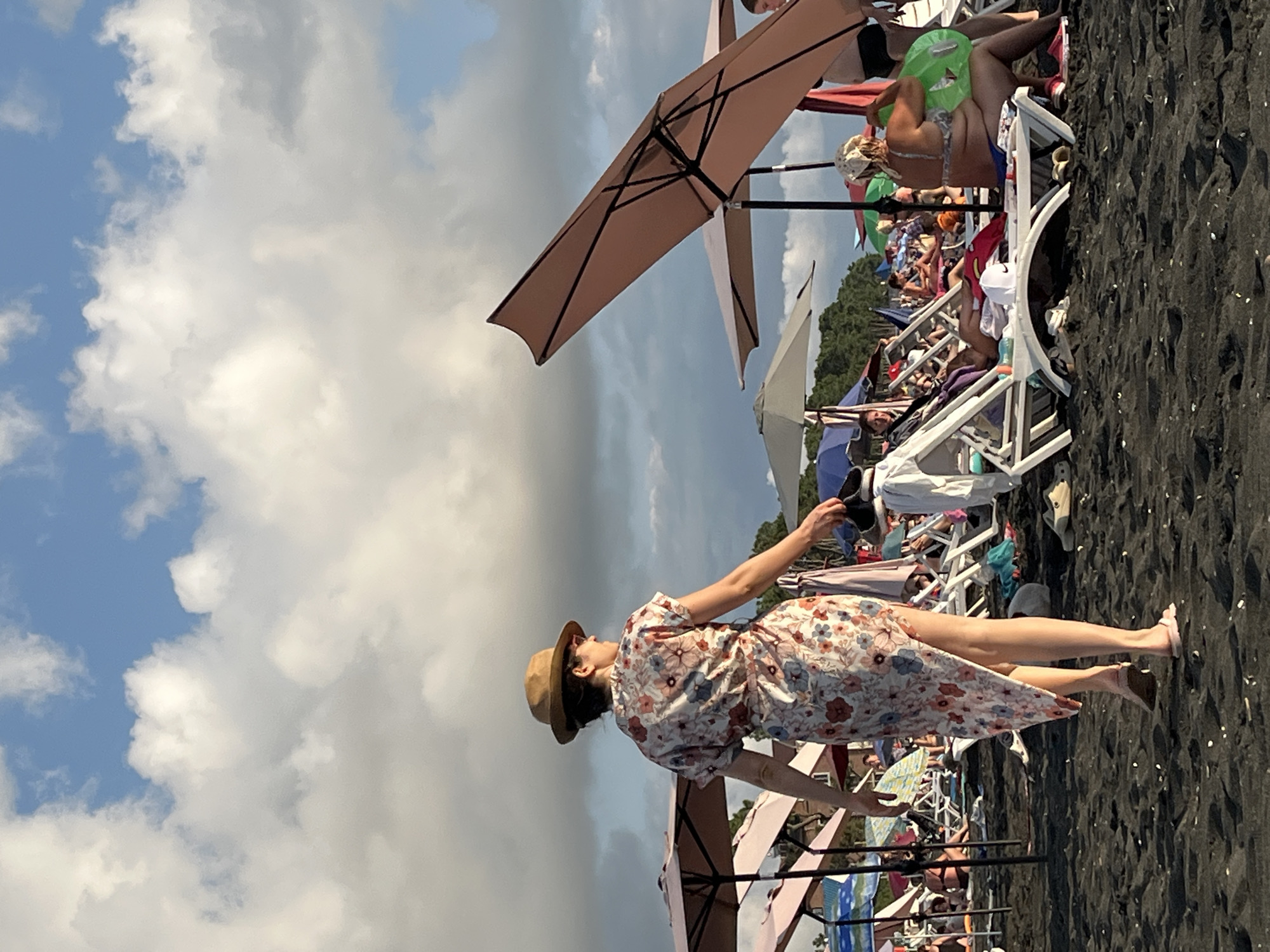 The image depicts a beach scene in Ureki, Georgia, characterized by its black sand. The foreground is dominated by a woman walking away from the camera, wearing a floral dress and a hat, and carrying a pair of shoes. The background features a crowded beach with numerous people relaxing on lounge chairs under umbrellas. The sky is partly cloudy with a mix of blue sky and white and grey clouds. The overall impression is a typical summer day at a popular beach resort.