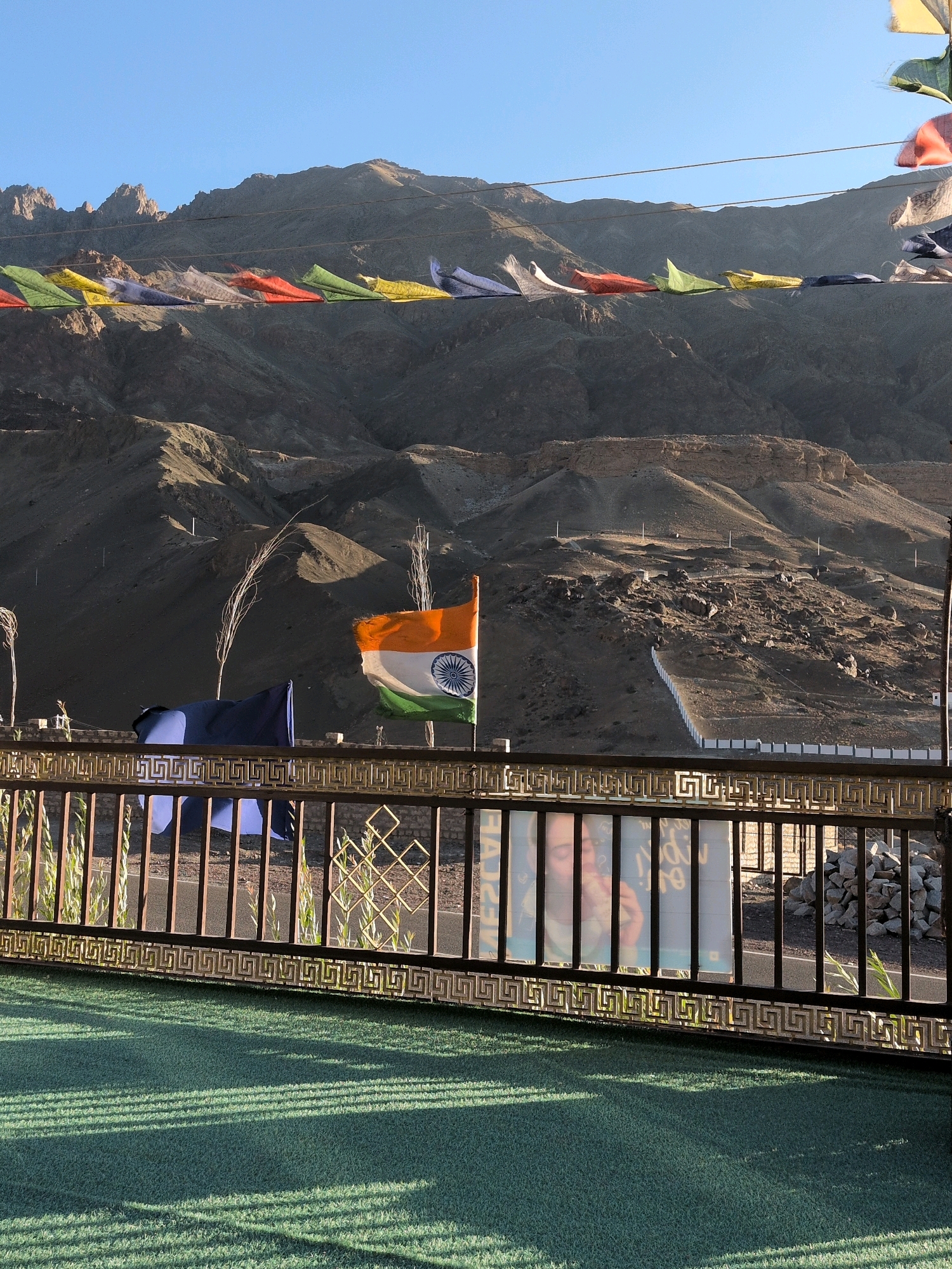The image depicts a mountainous landscape with prayer flags strung across the frame, in the city of Khaltse, India. In the foreground, an Indian flag is prominently displayed, waving in the breeze. A metal railing with a decorative geometric pattern separates the viewer from the background. Behind the railing, artificial green turf covers the ground, with distinct shadow patterns from the railing. The background consists of rugged, arid mountains and a clear blue sky. The overall scene suggests a high-altitude, possibly remote location.