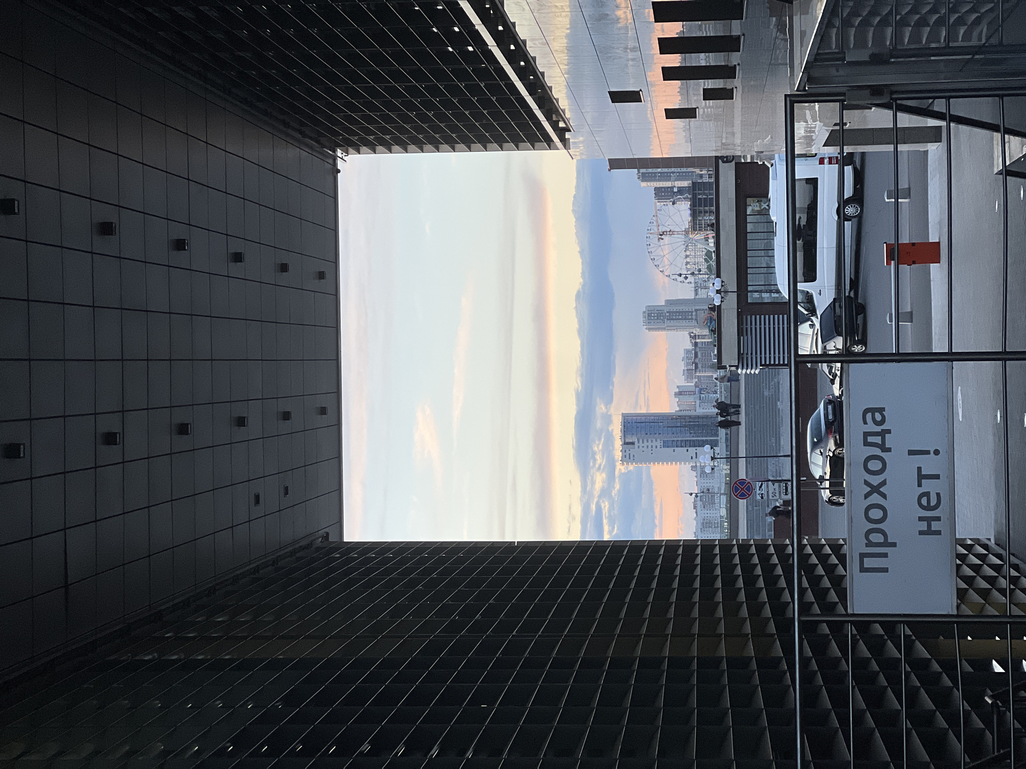 The image captures an urban scene in Kazan, Russia, framed by the architectural elements of two buildings with a textured, grid-like pattern. A view of the city opens between the buildings, revealing a ferris wheel, high-rise buildings, and a sky with hints of sunset colors. A white van and other cars are parked on the right side of the frame, near a sign with Russian text. People are faintly visible in the background. The perspective is taken from ground level, looking upwards.