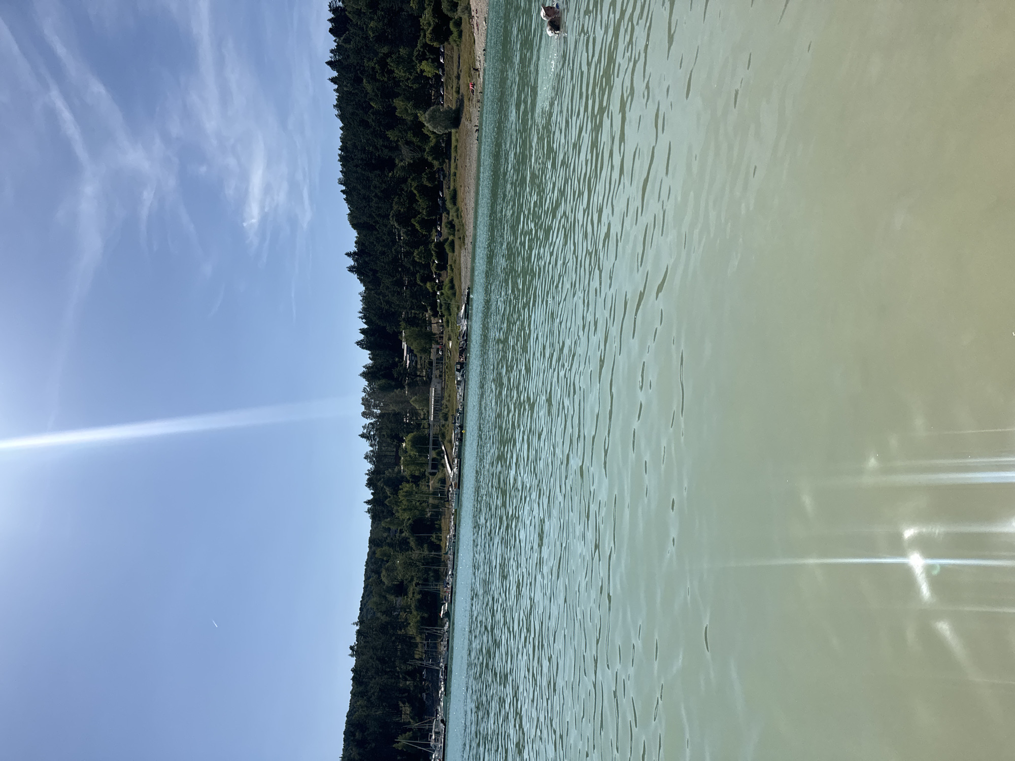 In Maisod, France, on a clear day, the calm, turquoise waters of a lake stretch towards a tree-lined shore. A dense forest covers the rolling hills in the background, with a few buildings and a road visible through the trees. Along the shore, a small beach is dotted with people enjoying the sun. Several sailboats are moored in the lake near the shore. In the foreground, two people are swimming in the water. The sun shines brightly, casting shimmering reflections on the water's surface.