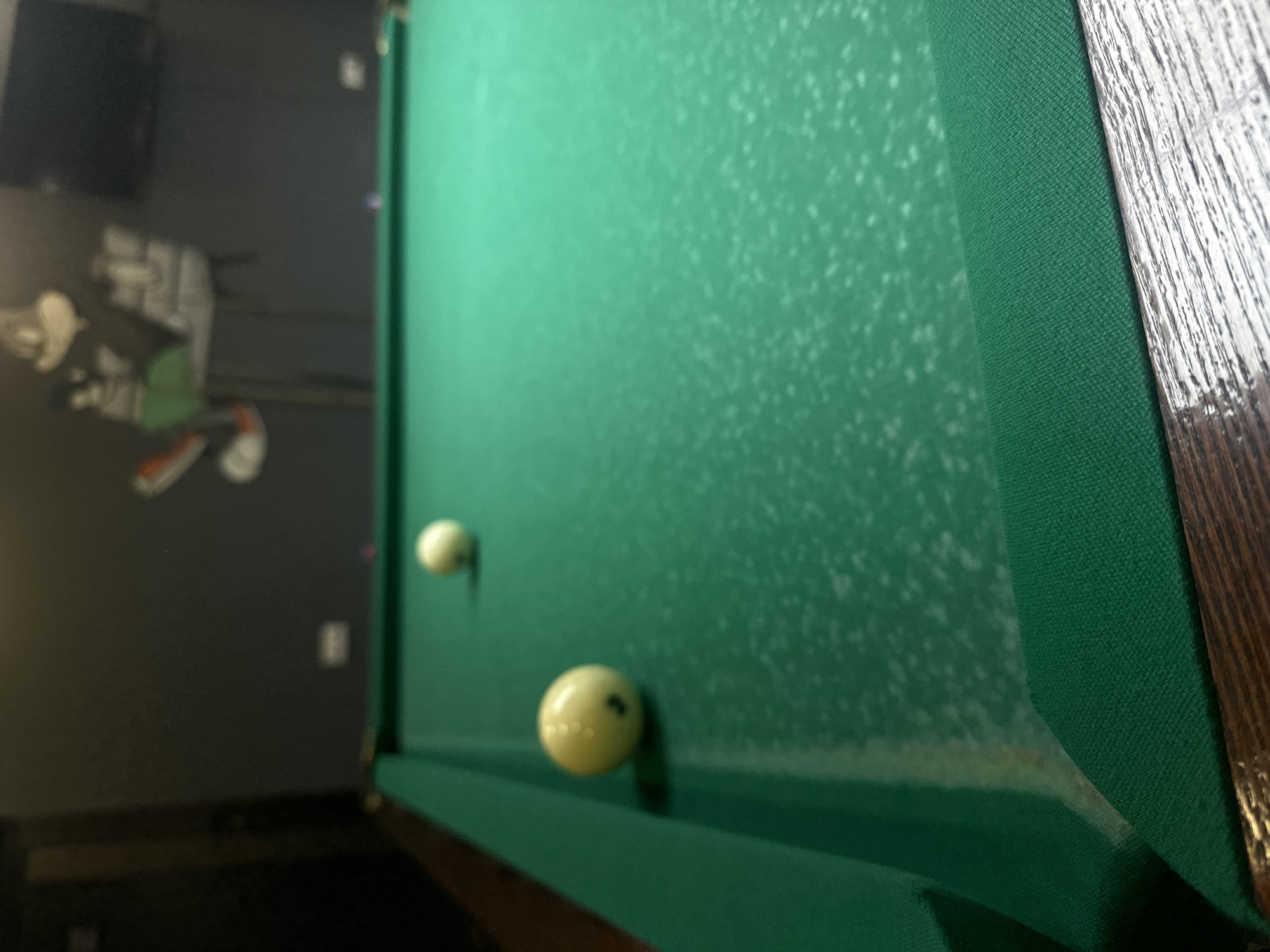 The image depicts a pool table covered in green felt. Two billiard balls, both white, are visible on the table. The table is positioned in a room with dark gray walls. A mural-like artwork is visible on the wall. The pool table has wooden trim. It cannot be determined if this is in Yerevan, Armenia.