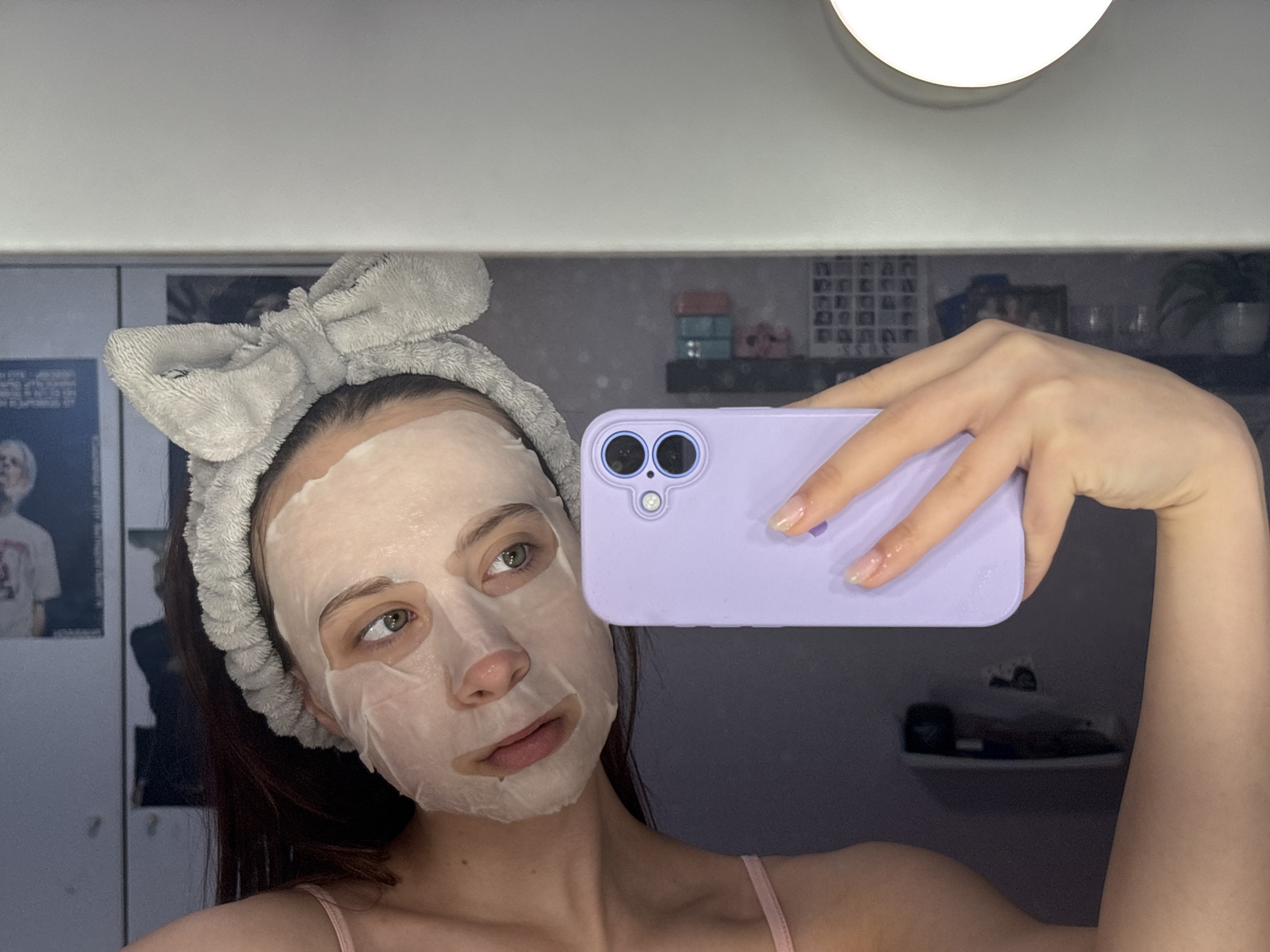 A person is taking a selfie in a bathroom. The person is wearing a white facial sheet mask and a gray headband with a bow on top. They are holding a light purple phone to take the photo. A round light fixture is visible in the upper right corner of the image. Behind the person, there is a mirror reflecting parts of the room, including shelves with items and a poster. The location is a bathroom, but it is impossible to determine the city of Прикамский, Russia from the provided image.
