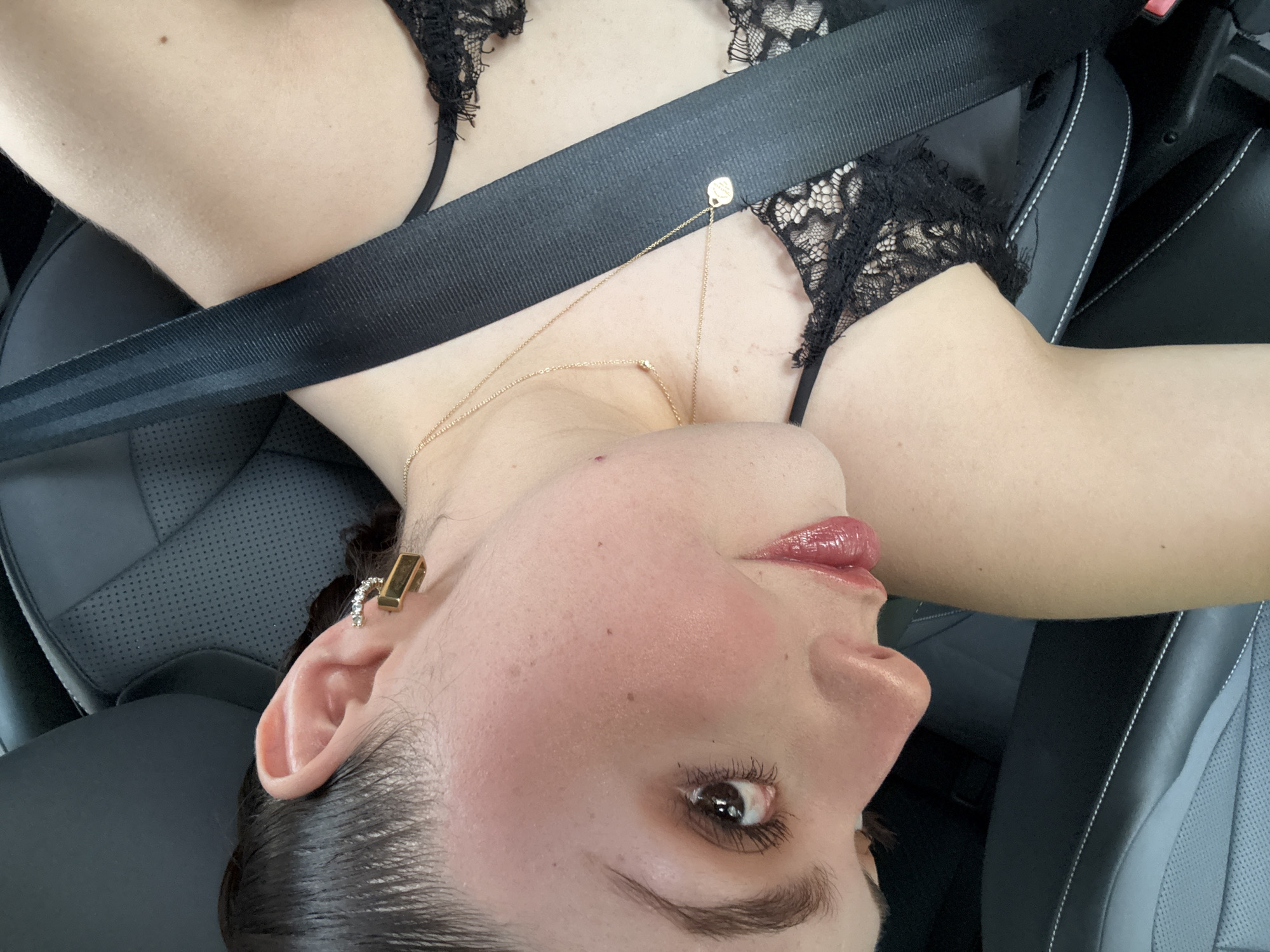 A woman with fair skin and brown hair is in a car, wearing a black lace top, a gold necklace, and earrings. She is wearing a seatbelt. The car's interior is black leather. The location is unknown and cannot be determined from the image.