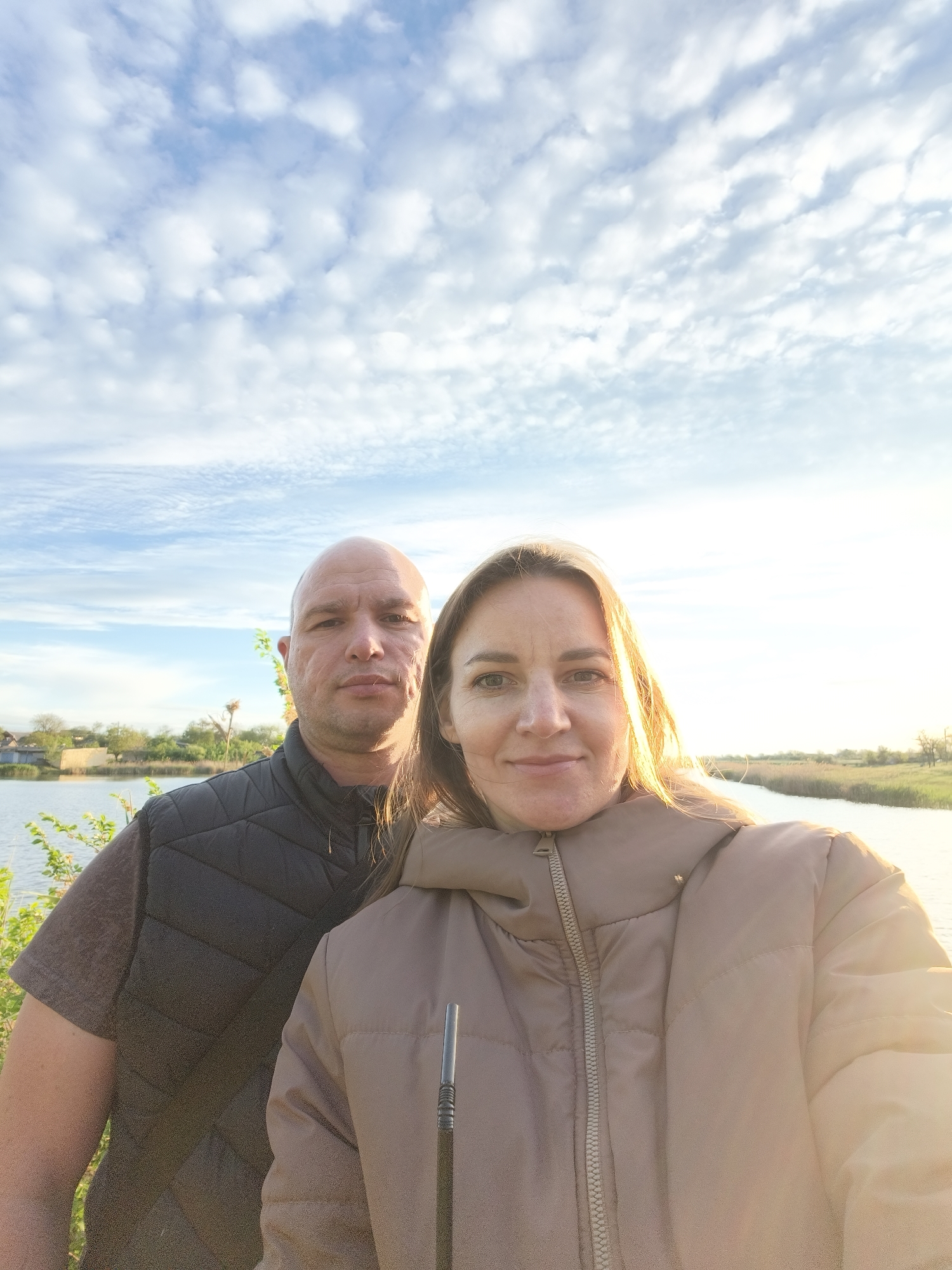 The image shows two people, a man and a woman, in an outdoor setting. The man is on the left with a shaved head, wearing a dark vest over a t-shirt. The woman is on the right, wearing a tan puffer jacket. Behind them is a body of water, likely a river or lake, with greenery along the banks. Buildings and trees are visible in the background. The sky is blue with scattered clouds. The image seems to have been taken in Shyroka Dacha, Ukraine.