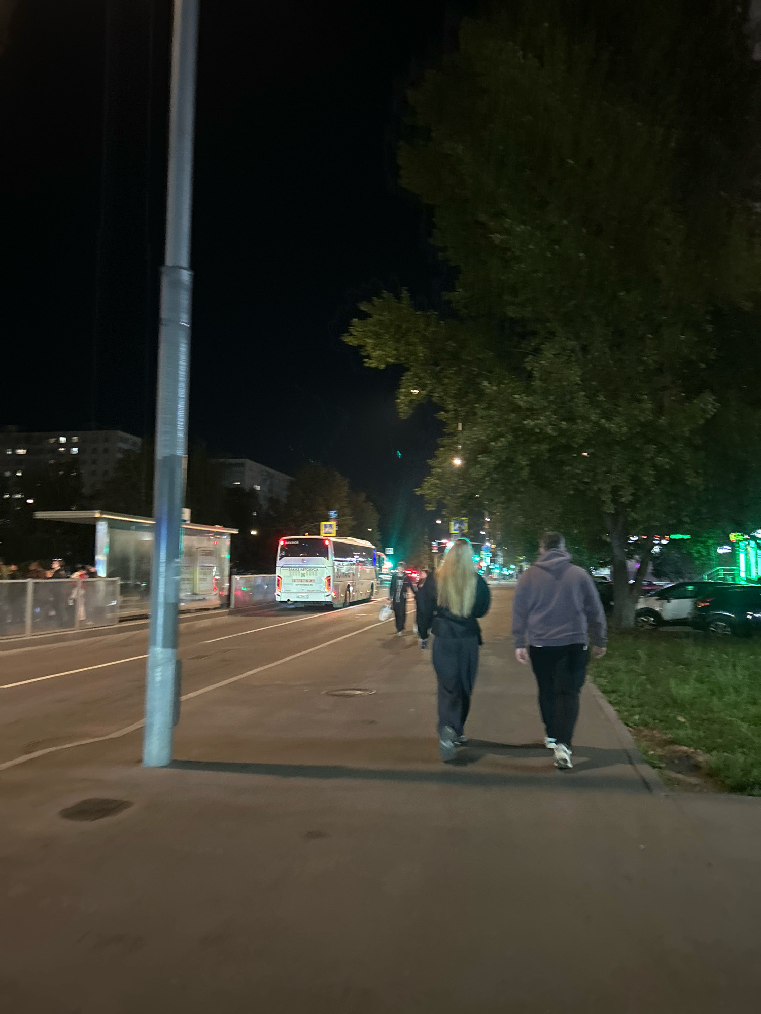 This is a night-time, eye-level, medium shot of a sidewalk scene on a street in Moscow, Russia. A tall, grey lamppost is positioned on the left side of the frame, casting a shadow. In the background, a white bus with red taillights is driving on the road. To the left of the bus is a bus stop with a glass-enclosed shelter. Several people are visible at the bus stop. In the foreground, two individuals are walking away from the camera on the sidewalk. The person on the left has long blonde hair and is wearing a black top and dark pants. The person on the right is wearing a purple hoodie and dark pants. Trees line the right side of the sidewalk, and parked cars are visible beyond the grass. The street is lit by artificial lights.