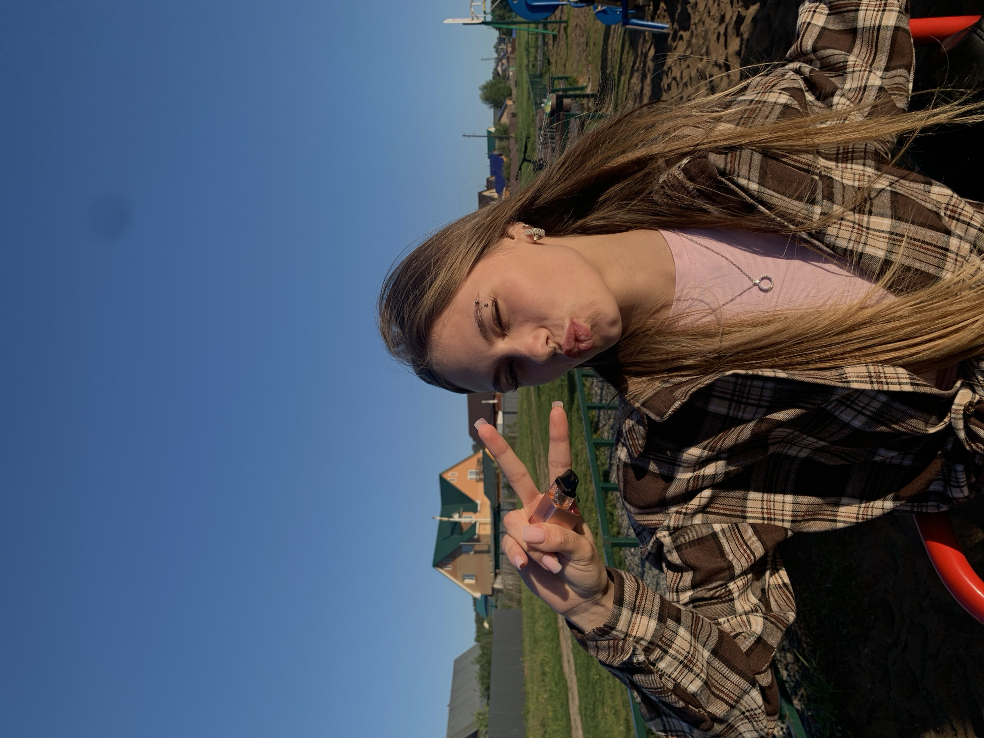 A young woman is pictured on a playground in Фоки, Russia, as the background contains residential buildings, possibly houses. The photo is taken from a low angle and tilted counterclockwise. The woman is posing with her eyes closed and lips pursed, holding up a peace sign. In her fingers, she is holding a small perfume bottle. She is wearing a light pink top under a brown and white plaid shirt, and a silver necklace with a circular pendant. She has a piercing above her eyebrow. Her long, blonde hair is draped across her chest and shoulder. The background consists of a clear blue sky and some playground equipment. There is also a house behind her.