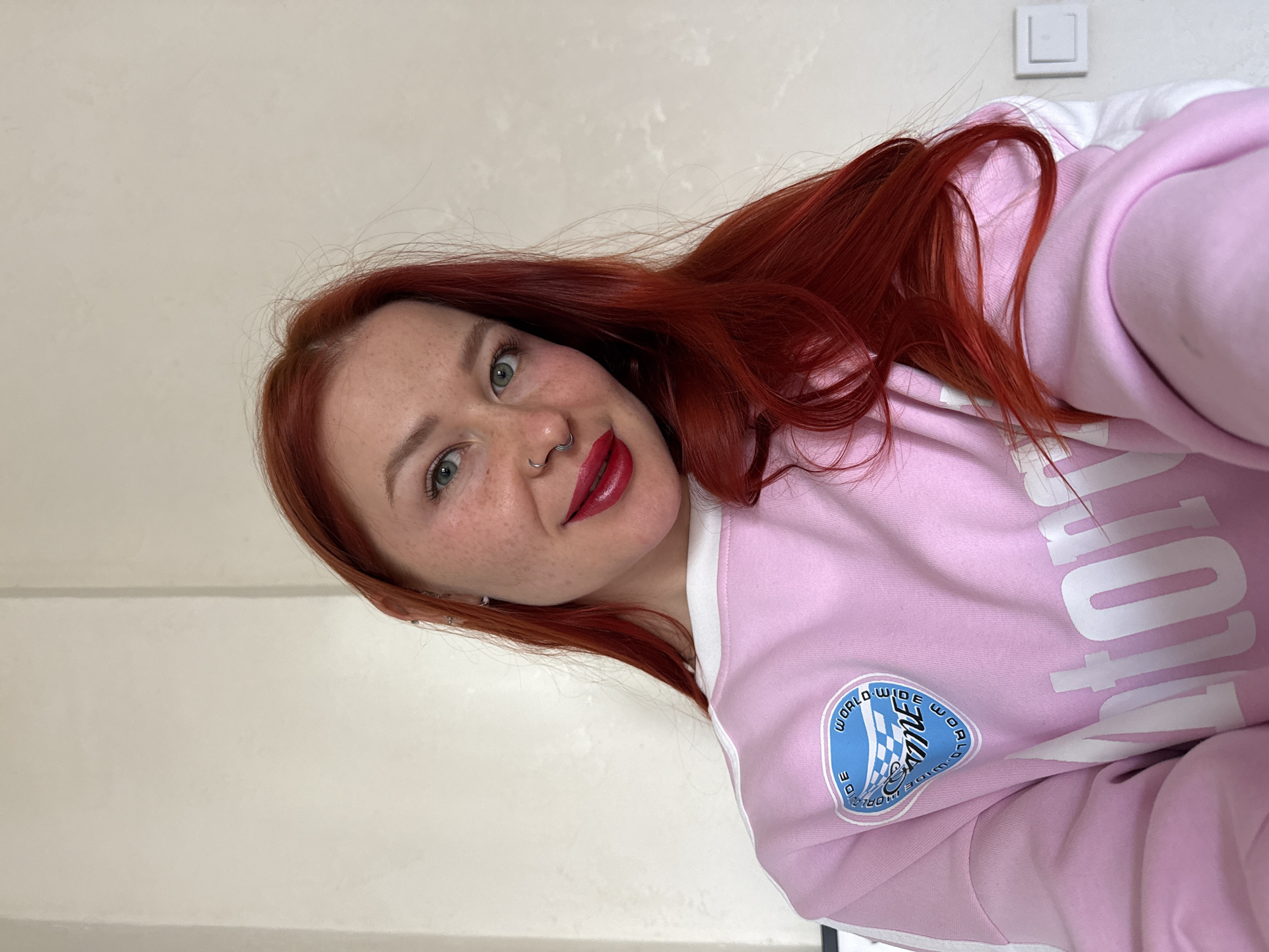 An indoor selfie captures a young woman with vibrant red hair, green eyes, and noticeable freckles across her face. She has a septum piercing and is wearing bright red lipstick, slightly smiling as she looks directly at the camera. Her hair is long and falls around her shoulders. She is dressed in a light pink hoodie or sweatshirt with white trim along the shoulders and arms. On her right arm, there is a circular patch with a blue background featuring a checkered flag design and the text "WORLDWIDE" encircling it. Partial white text, appearing to read "OUT ON," is visible on the front of her hoodie. The background is a plain, light-colored wall, with a white light switch visible in the upper right corner of the frame. The lighting suggests it is daytime. The image was taken in Rostov-on-Don, Russia.