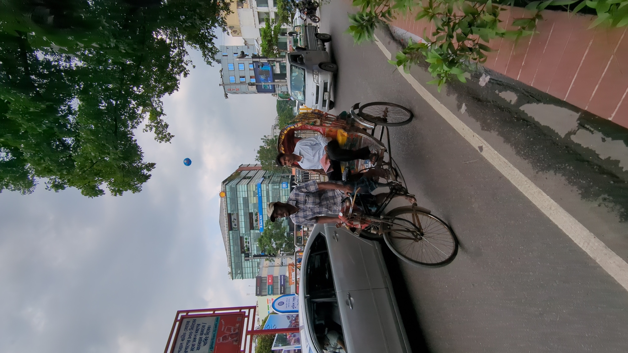 The image shows a rickshaw carrying a passenger in Dhaka, Bangladesh. The rickshaw is on a street with cars and other vehicles. A sign in the foreground reads "গন্যাল ব্যতীত মোড়ের রপাশের ১০০ মিটারের মধ্যে সকল ধরণের যানবাহন থামানো / পার্কিং নিষেধ।" (translates to "No stopping/parking of any kind of vehicles within 100 meters of the signal except on the corner"). There are buildings in the background, including one with the "Bank Asia" logo. The sky is overcast. A blue balloon is visible above the trees.