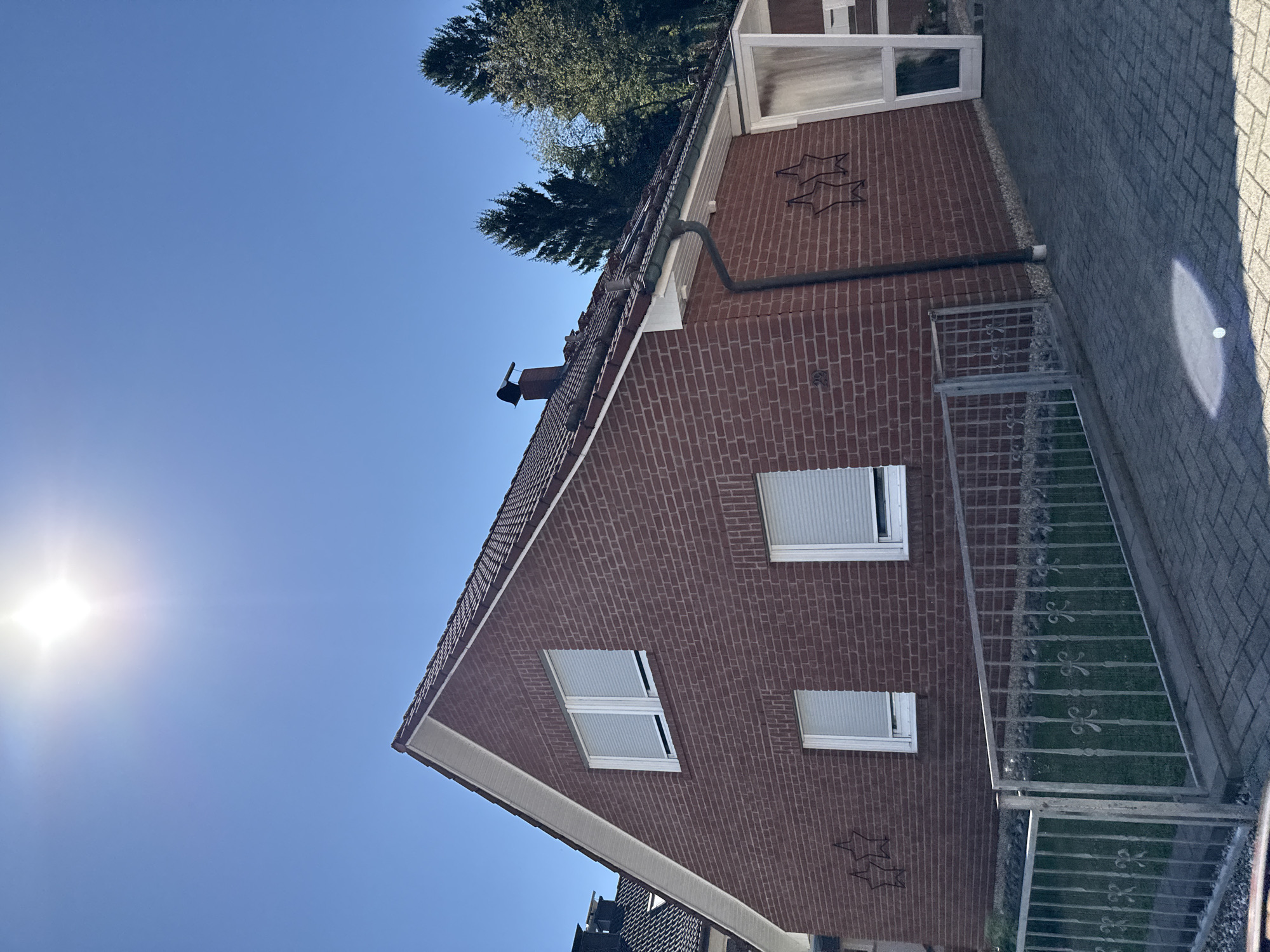 The image captures a side view of a two-story brick house on a sunny day in Bad Lauterberg im Harz, Germany. The house has a red tile roof, white framed windows with closed blinds, and a prominent chimney. A metal fence encloses a small lawn area in front of the house, and the driveway is paved with interlocking bricks. Two star-shaped decorations are affixed to the brick wall. In the background, lush green trees are visible against a clear blue sky, and the sun is shining brightly, casting a glare on the scene.