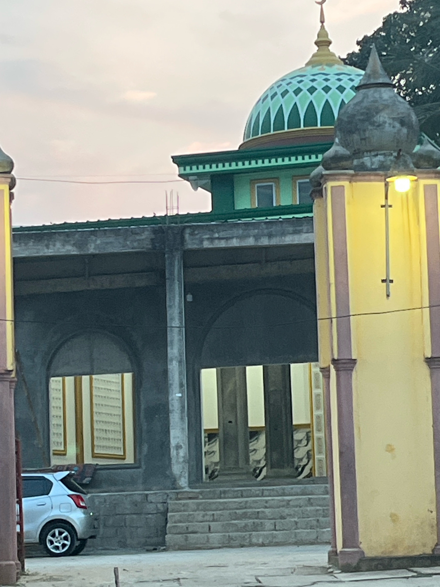 The image shows a partially constructed mosque in Kebumen, Indonesia. The main structure has a green dome with a crescent moon finial and is surrounded by construction scaffolding. In the foreground, a yellow minaret-like structure with dark red trim stands to the right, and a similar structure is partially visible on the left. A silver car is parked to the left of the building, near a set of stone steps leading up to an arched entryway. The sky is overcast and appears to be either dawn or dusk. The scene suggests an ongoing construction project or renovation.