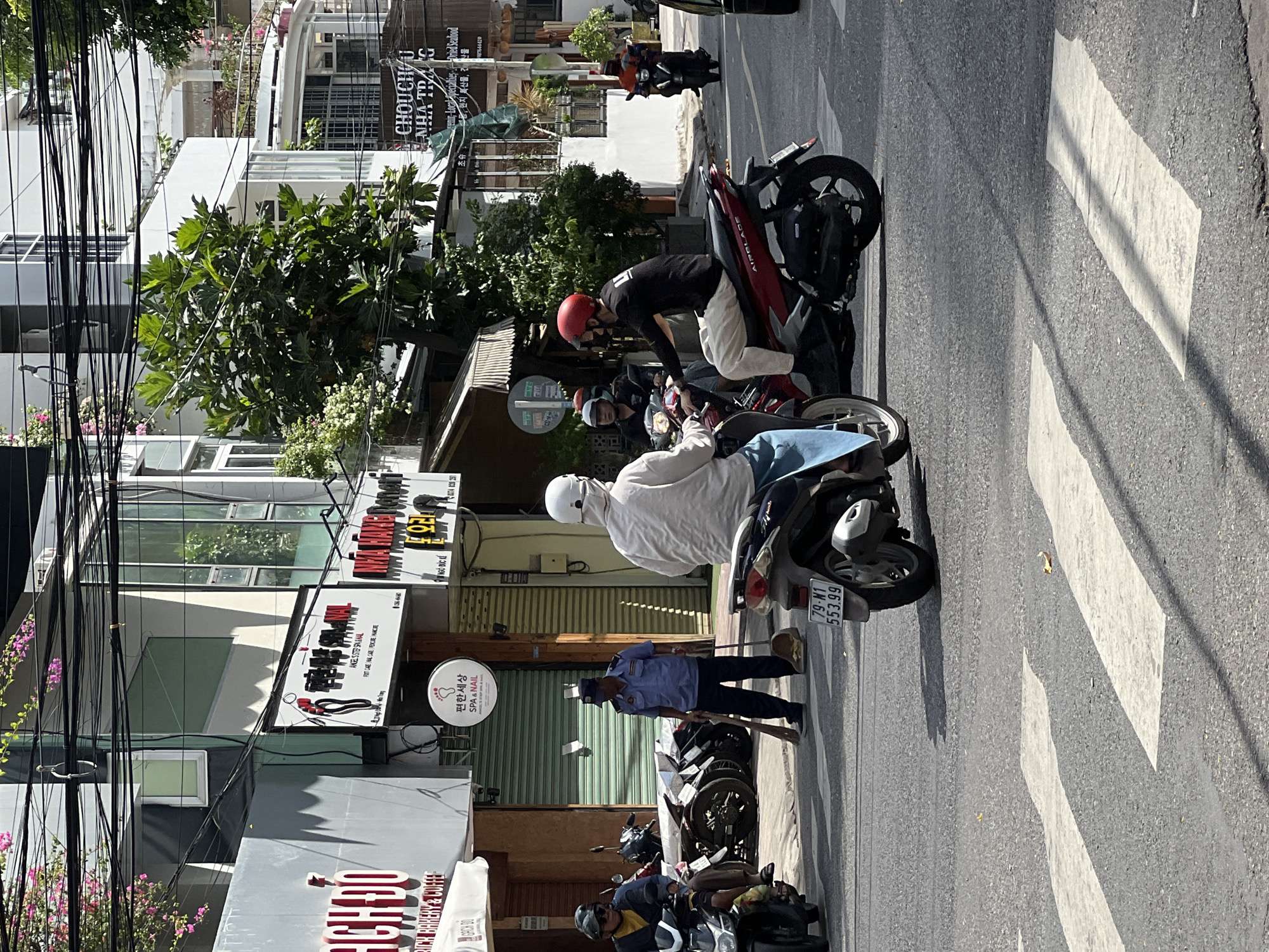 The image depicts a street scene in Nha Trang, Vietnam, likely near a commercial area, given the presence of storefronts with various signage. In the foreground, a crosswalk is visible on the asphalt road. Several motorcycles are present, with two prominent ones in the center. On the left, a person wearing a yellow shirt and dark helmet is on a black motorcycle. To their right, a person in a white long-sleeved shirt and light blue pants is on a red motorcycle. This rider is wearing a white helmet that covers their face. Behind them, on another red motorcycle, a person wearing a black long-sleeved shirt and light-colored pants is also on a motorcycle, wearing a red helmet.
In the middle ground, a man in a blue uniform and cap is holding a broom and dustpan, appearing to be cleaning the street. He is positioned between parked motorcycles on the sidewalk. The buildings lining the street feature multiple stories, with balconies and windows visible. Signage in various languages, including Vietnamese and what appears to be Korean, indicates businesses such as a bakery and coffee shop, a spa, and a restaurant or food establishment. Overhanging the street is a dense network of electrical wires, a common sight in urban Vietnam. The overall lighting suggests it is daytime with strong sunlight, casting distinct shadows on the road.