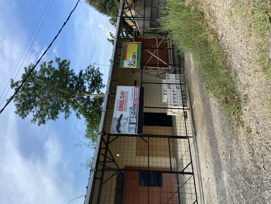 The image shows a storefront located at coordinates -6.643, 107.417 in the city of Sukatani, Indonesia. The storefront is a single-story building with a black metal gate and a red brick facade. It has a sign on the front advertising "UNILON", a brand of plumbing pipes. There is also a sign advertising "Romatex", likely a building material. The sign is mounted on a wooden frame. There is a large, green tree to the left of the building. White cinder blocks are stacked in front of the storefront. The scene is taken from across the street, looking towards the storefront. There are no people in the image. The ground is a gravel path. There is green grass in the foreground. The sky is blue with a few white clouds. The image is likely taken during the day.