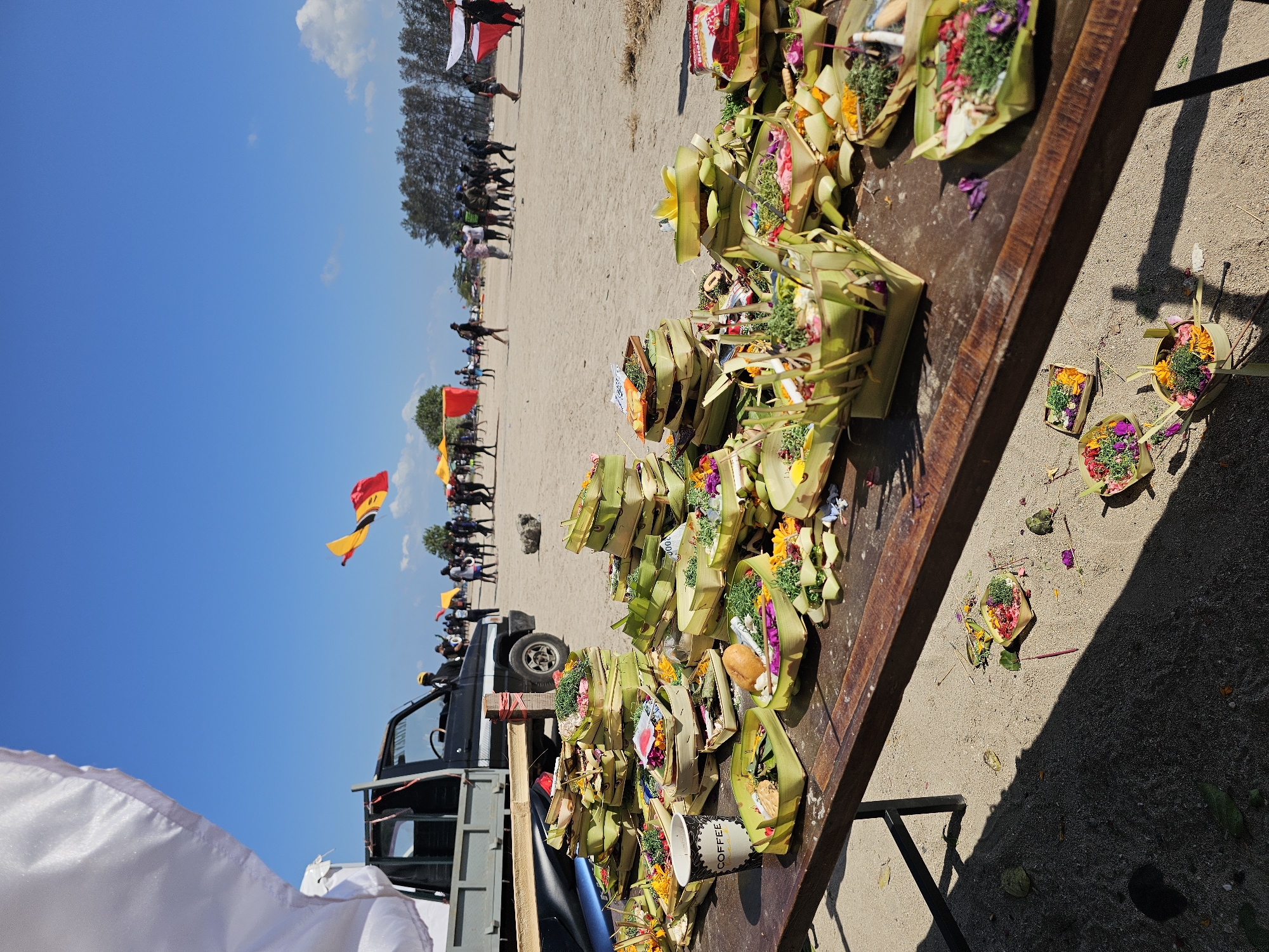 This image shows a beach scene in Denpasar, Indonesia, likely during a cultural event involving offerings. In the foreground, a wooden table is laden with numerous small, woven palm leaf baskets filled with colorful flowers, greenery, and what appear to be food items, suggesting offerings or ceremonial items. A paper cup with the word "COFFEE" is also present on the table. A black pickup truck is parked behind the table, and a white flag is partially visible to the left. In the background, people are gathered on the sandy beach, some holding or observing kites flying in the bright blue sky. Trees line the horizon. The overall impression is of a lively outdoor gathering with a focus on cultural traditions and celebration.