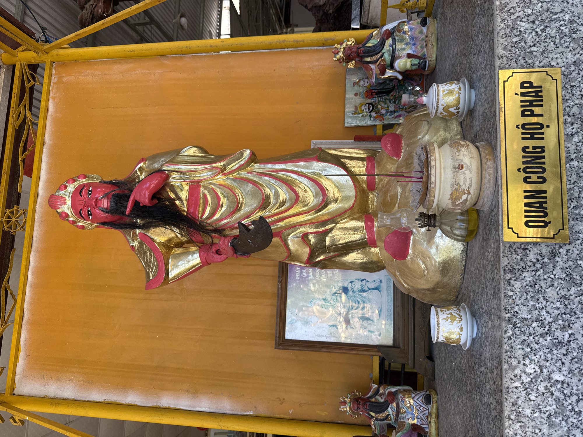 This image depicts a shrine in Bac Nha Trang Ward, Vietnam, dedicated to Quan Cong, a revered Chinese general and deity in Vietnamese folk religion. The central figure is a large, striking statue of Quan Cong, painted a vibrant red and adorned in a shimmering gold and red robe, featuring a black beard and an elaborate headdress. The statue stands on a circular, golden platform, with its right hand held up in a commanding gesture.
In front of the statue, an altar is set up with various religious items. These include a framed picture displaying a seated figure, several small ceramic cups and a lidded jar decorated with golden dragons, an incense burner with several red incense sticks, and a small oil lamp with a glass reservoir. Flanking the central statue are smaller figurines of deities or attendants, also richly decorated.
A granite countertop forms the base of the shrine, on which the altar items are placed. A prominent golden plaque with black text, reading "QUAN CÔNG HỘ PHÁP" (Quan Cong the Dharma Protector), is affixed to the front of the countertop, identifying the dedication of the shrine. The background is a large, orange-yellow wall, possibly part of a larger structure, with a yellow metal frame extending from the top and sides, suggesting an outdoor or semi-outdoor setting. The overall atmosphere is one of reverence and devotion, typical of such religious sites.
