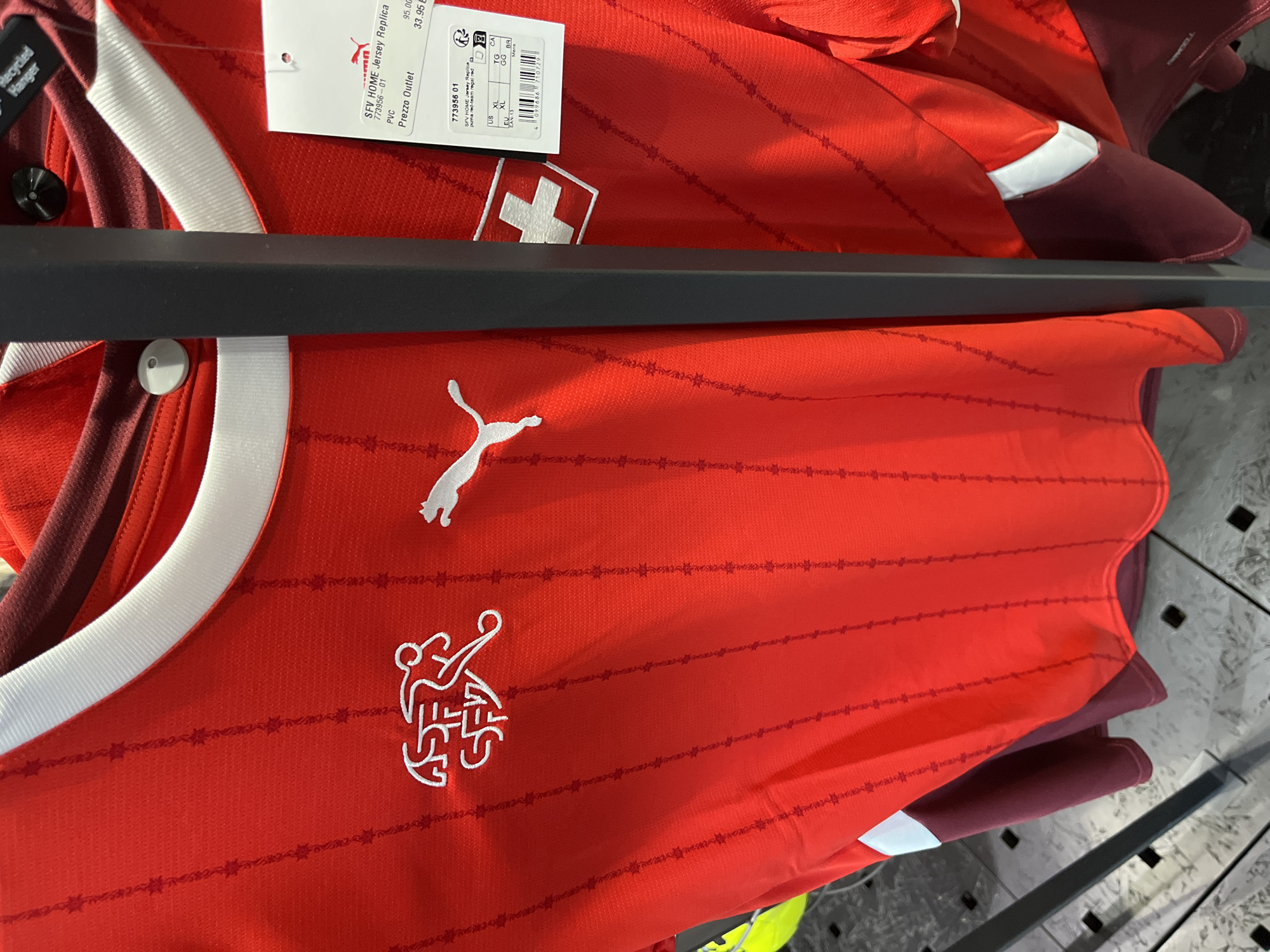 A red Puma Switzerland national football team jersey is displayed on a hanger in a retail store. The jersey features a white crew neck collar and white Puma logos on the chest. The Swiss Football Association (SFA) crest is also visible on the left chest. The jersey is made of a textured fabric with a subtle vertical stripe pattern. A price tag is attached to the right side of the jersey, displaying product information and pricing in Euros. The background shows other clothing items and display racks, typical of a sports apparel store. The store is located in Rodengo, Italy.