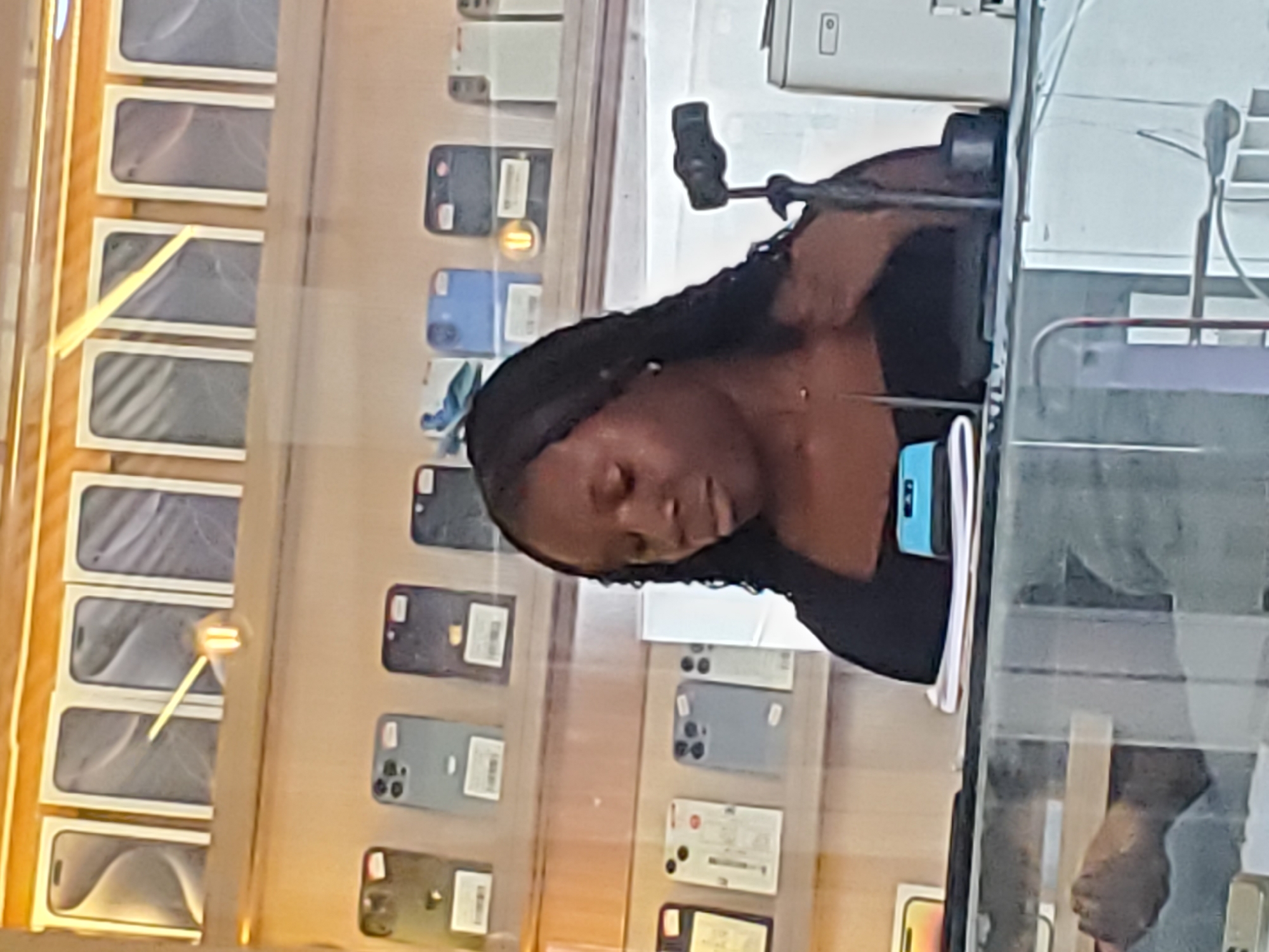The media file shows a young Black woman with braided hair, wearing a black top, leaning over a counter in what appears to be an electronics store. She is looking down, possibly at a phone or other device, with her eyes closed. A camera rig is mounted on the counter in front of her, pointing towards her face. Reflected in the glass counter are shelves filled with cell phone boxes, some stacked horizontally and others vertically. The phone boxes are mostly white with images of phones on them. There are also individual phones displayed vertically on shelves to the right, showcasing various models and colors. The lighting suggests it could be daytime, but the indoor setting makes it difficult to determine the exact time of day or weather. The scene conveys a sense of quiet concentration or possibly fatigue on the part of the woman. The location cues suggest an electronics retail environment, likely a phone shop, possibly in Jos, Nigeria, as indicated by the context. No visible text is legible within the image.