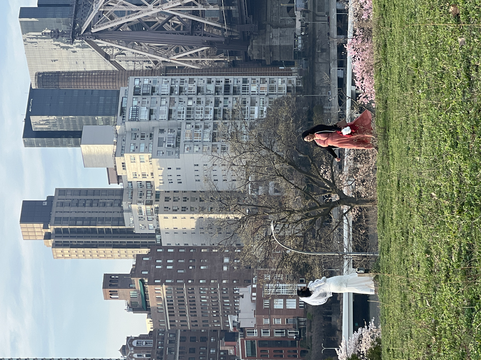 This photograph captures an outdoor scene in New York City. The background features a dense urban landscape of high-rise buildings and infrastructure, including portions of a bridge structure. Two individuals are present; one is wearing a long, white dress and possibly a veil, while the other wears a reddish-brown dress. They are positioned in a green space, possibly a park, with grass and flowering trees. The image's orientation is off, potentially due to the camera's perspective.