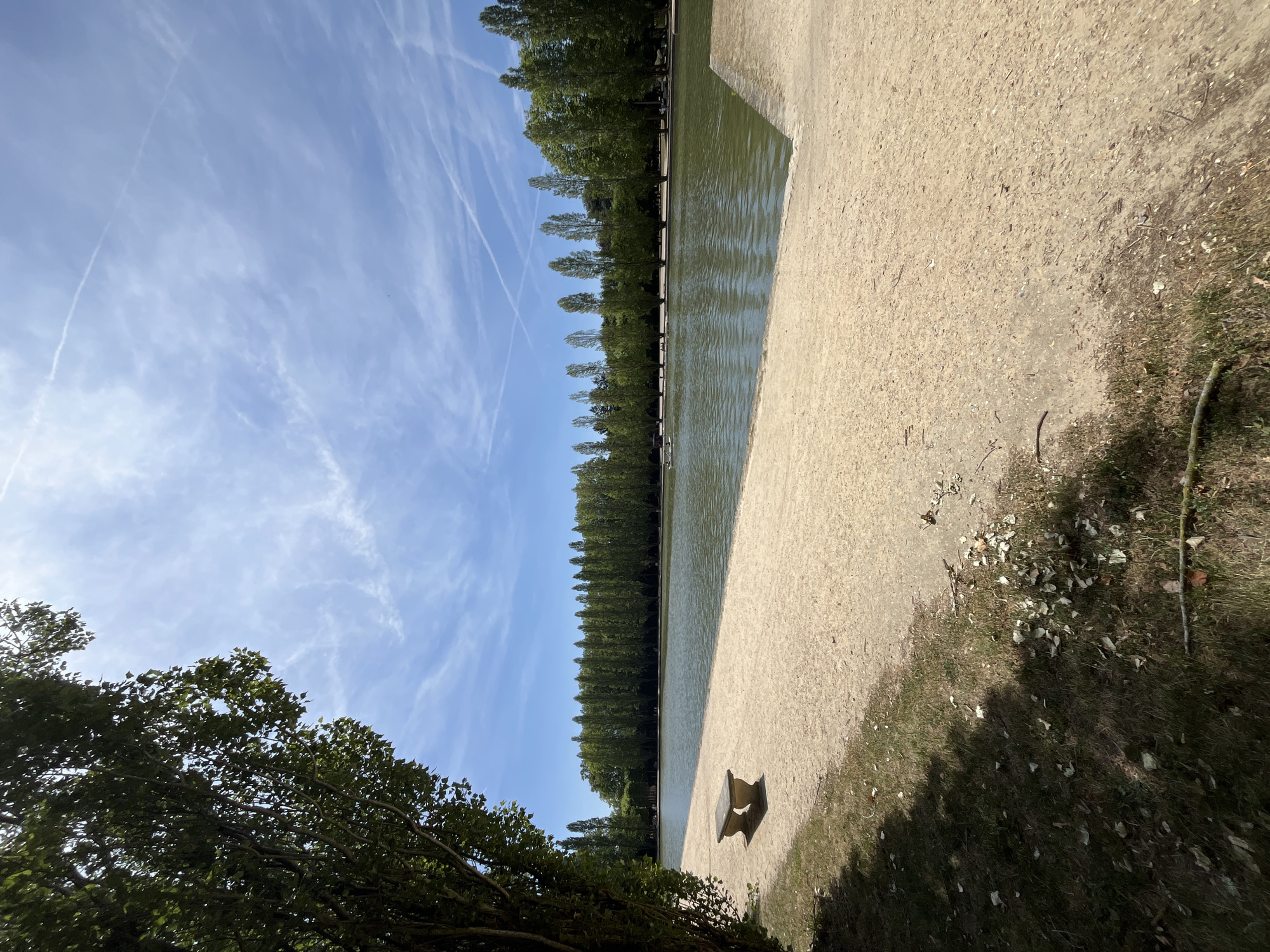 The image depicts a tranquil scene at the Parc d'Est dans la ville d'Antony, France. A large, calm body of water occupies the central portion of the frame, bordered by a sandy bank in the foreground and a manicured line of tall, slender trees in the background. A solitary stone bench sits on the sand to the left, casting a shadow. The sky above is a clear blue, adorned with wispy white clouds and contrails from aircraft. Lush green foliage from trees frames the left side of the image, suggesting a park or garden setting. In the distance, on the water, a small boat with several occupants can be observed. The overall impression is one of a serene summer day in a well-maintained public park.