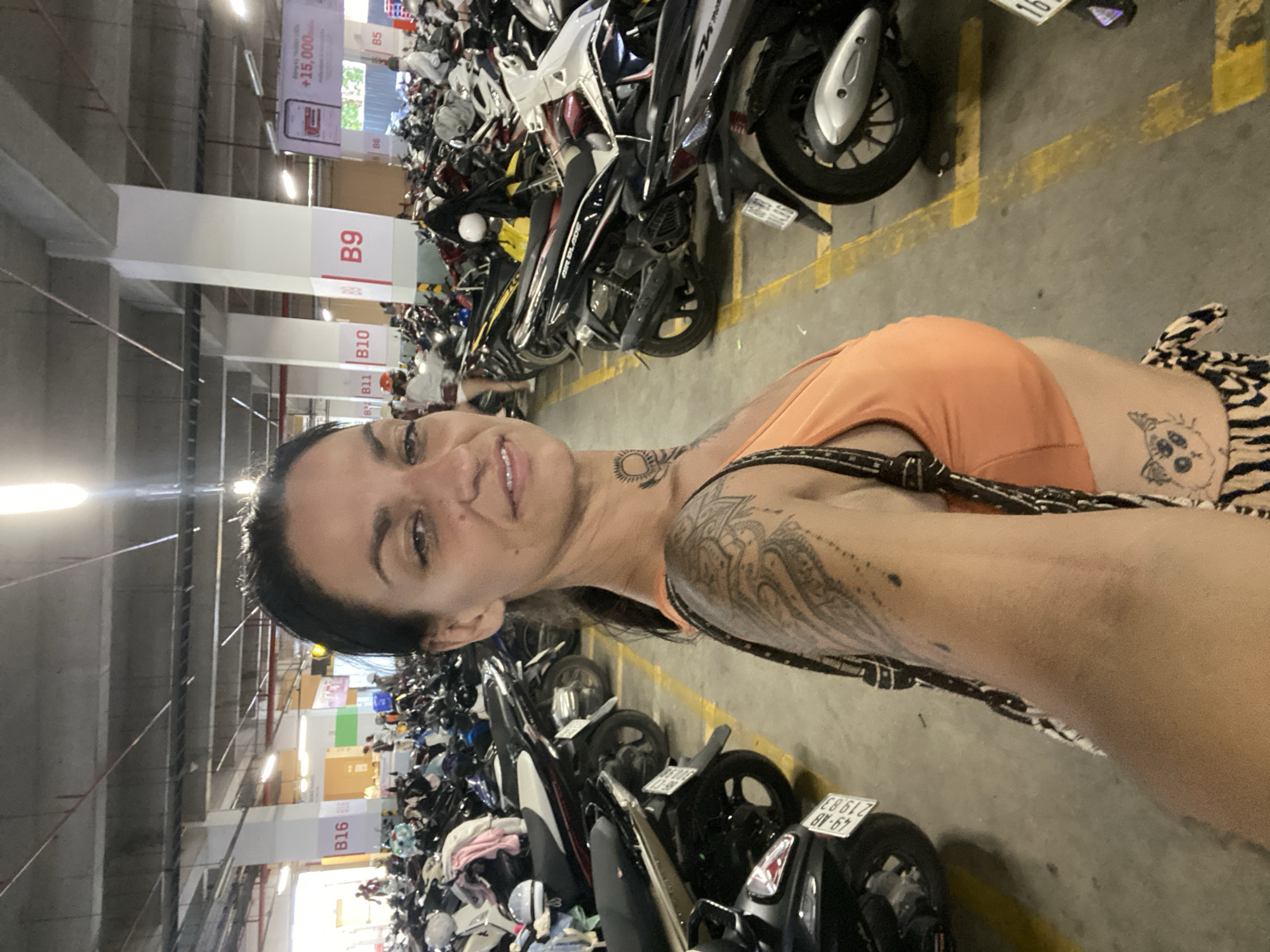 The image shows a woman in a parking garage. She is wearing an orange crop top and a patterned skirt. She has tattoos on her arm and neck. The parking garage is filled with motorcycles. There are signs in Vietnamese on the pillars. The overall scene suggests a busy urban environment. The location is identified as Phu Thuy Ward, Vietnam.