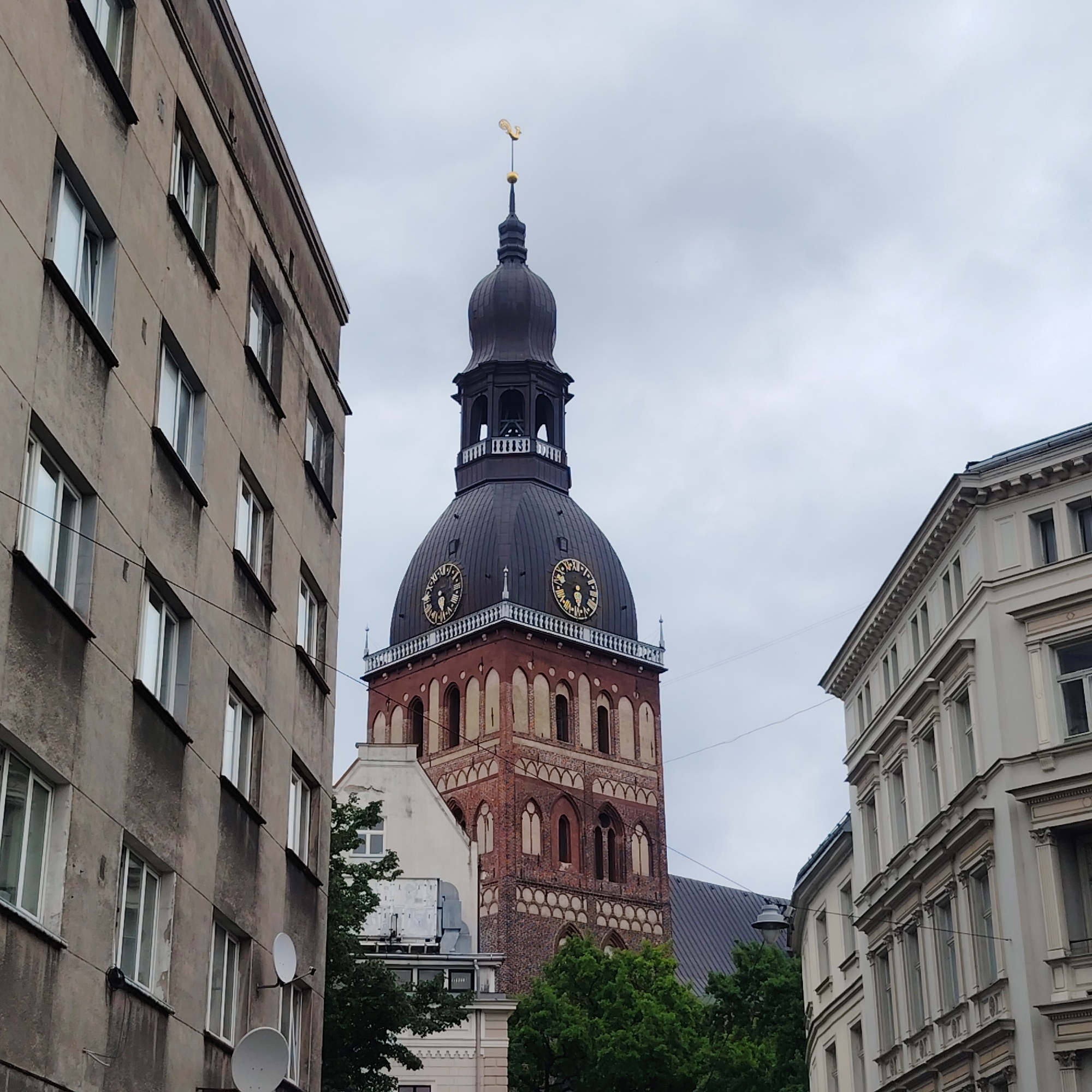 The image shows a view of the Riga Cathedral in Riga, Latvia, from a narrow street between two buildings. The cathedral's tall, dark spire dominates the background, topped with a golden weathervane. The spire is flanked by two large clock faces on its upper section. The foreground is occupied by the facades of two distinct buildings. On the left is a weathered, multi-story apartment building with rows of uniformly spaced windows. On the right, a more ornate, lighter-colored building with classical architectural details is visible. The sky is overcast, suggesting a cloudy day. The street appears to be a pedestrian area or a quiet street, as no vehicles or pedestrians are present. The overall impression is one of an urban landscape dominated by historical architecture.