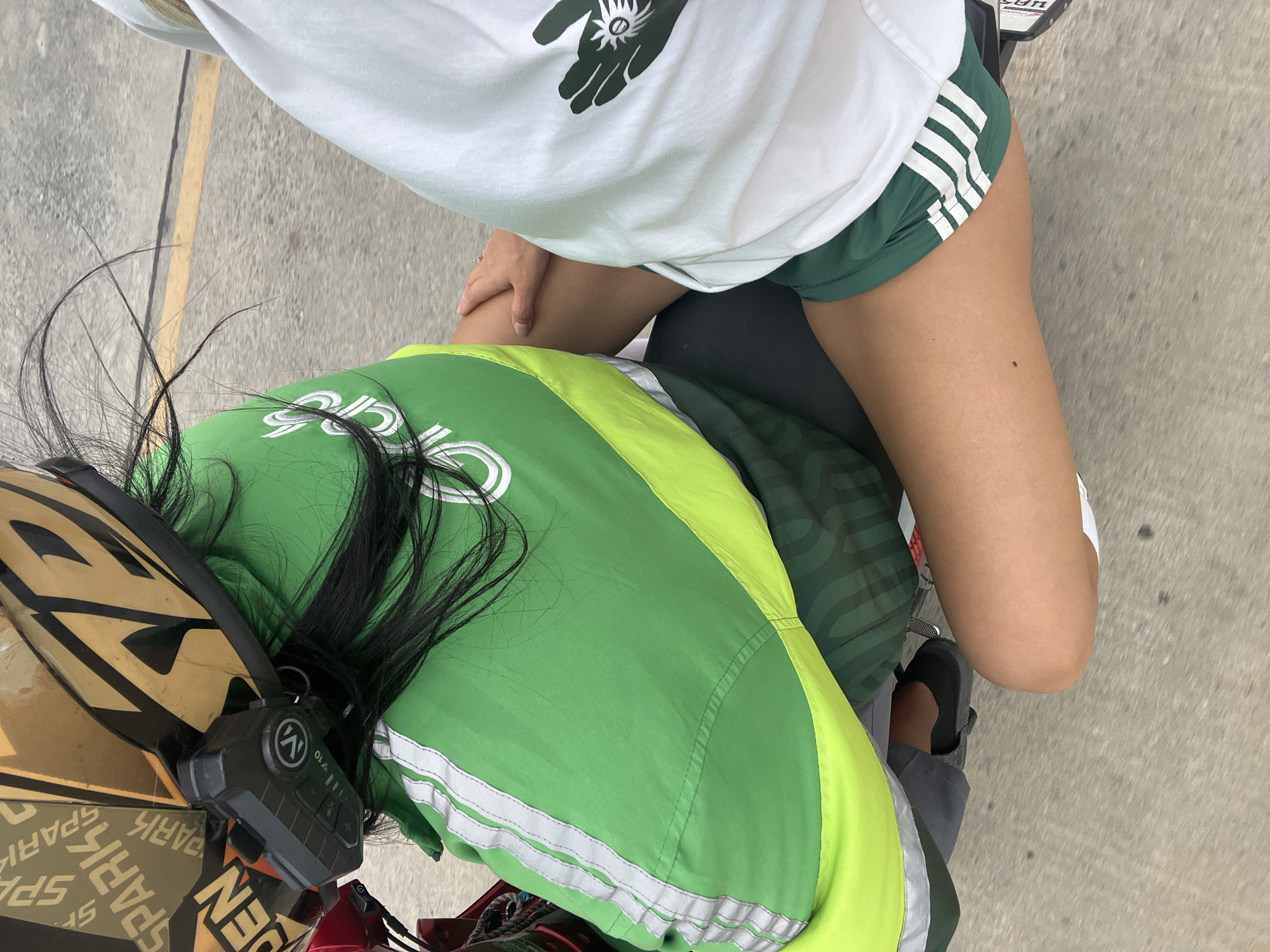 A high-angle shot depicts two individuals on a motorcycle or scooter on a paved surface, likely a street. The person in the foreground is wearing a green and yellow "Grab" branded jacket, a motorcycle helmet, and has long black hair. The helmet has a black communication device attached. The other individual, presumed to be a passenger, wears a white t-shirt with a stylized hand print, and green Adidas-style shorts with white stripes. One hand is visible on the rider's knee. The passenger's footwear consists of black sandals.