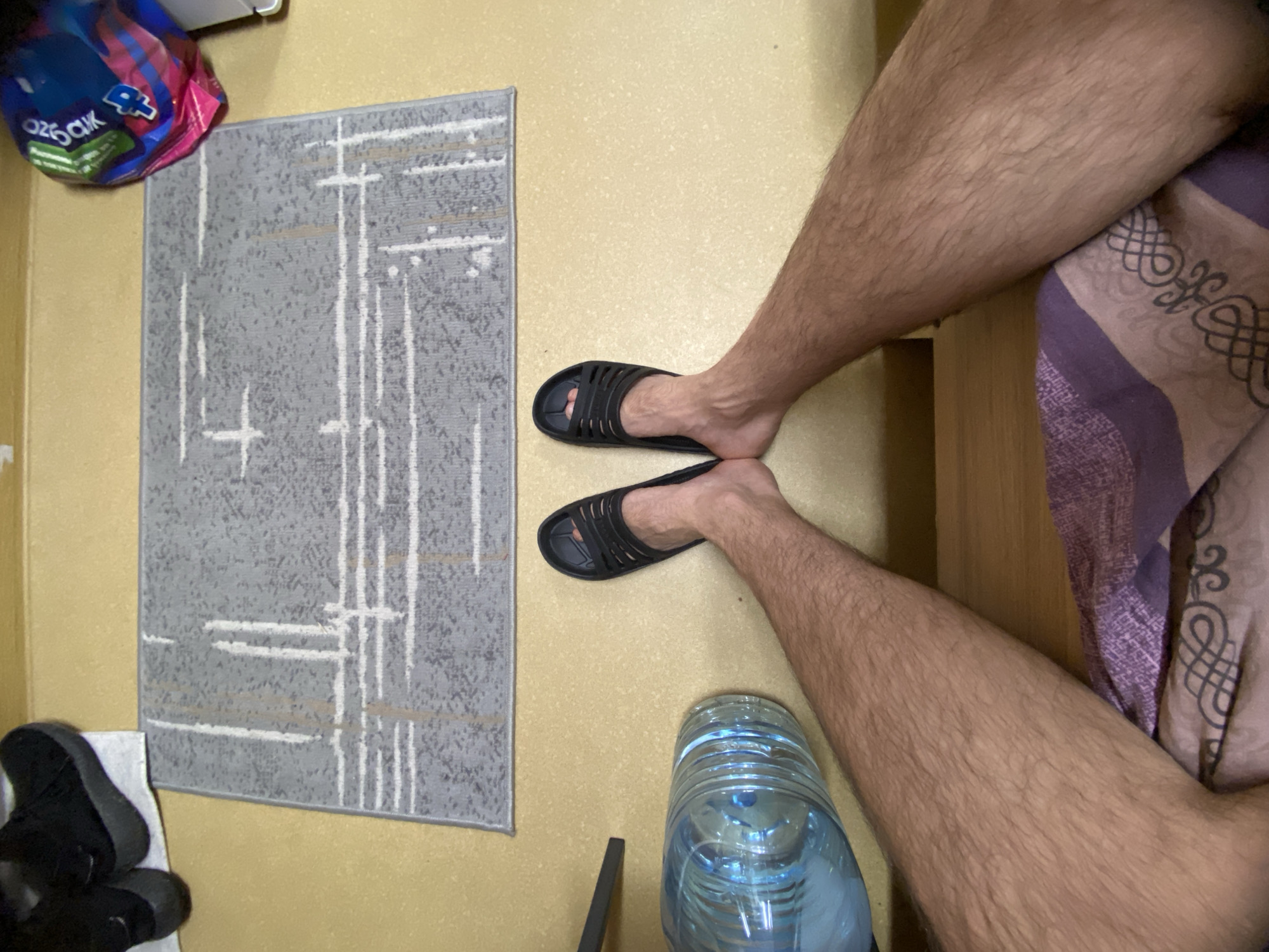 A high-angle shot reveals a section of what appears to be a room in Koskolovo, Russia. The floor is covered in light beige linoleum. A grey rug with a geometric pattern lies to the left. A pair of black rubber sandals sit on the linoleum. Two hairy legs are positioned centrally. Beside the legs is a blue plastic water bottle. Part of a bed covered with a patterned sheet is visible at the top. A bag is in the top left. A pair of black boots lies at the bottom left.