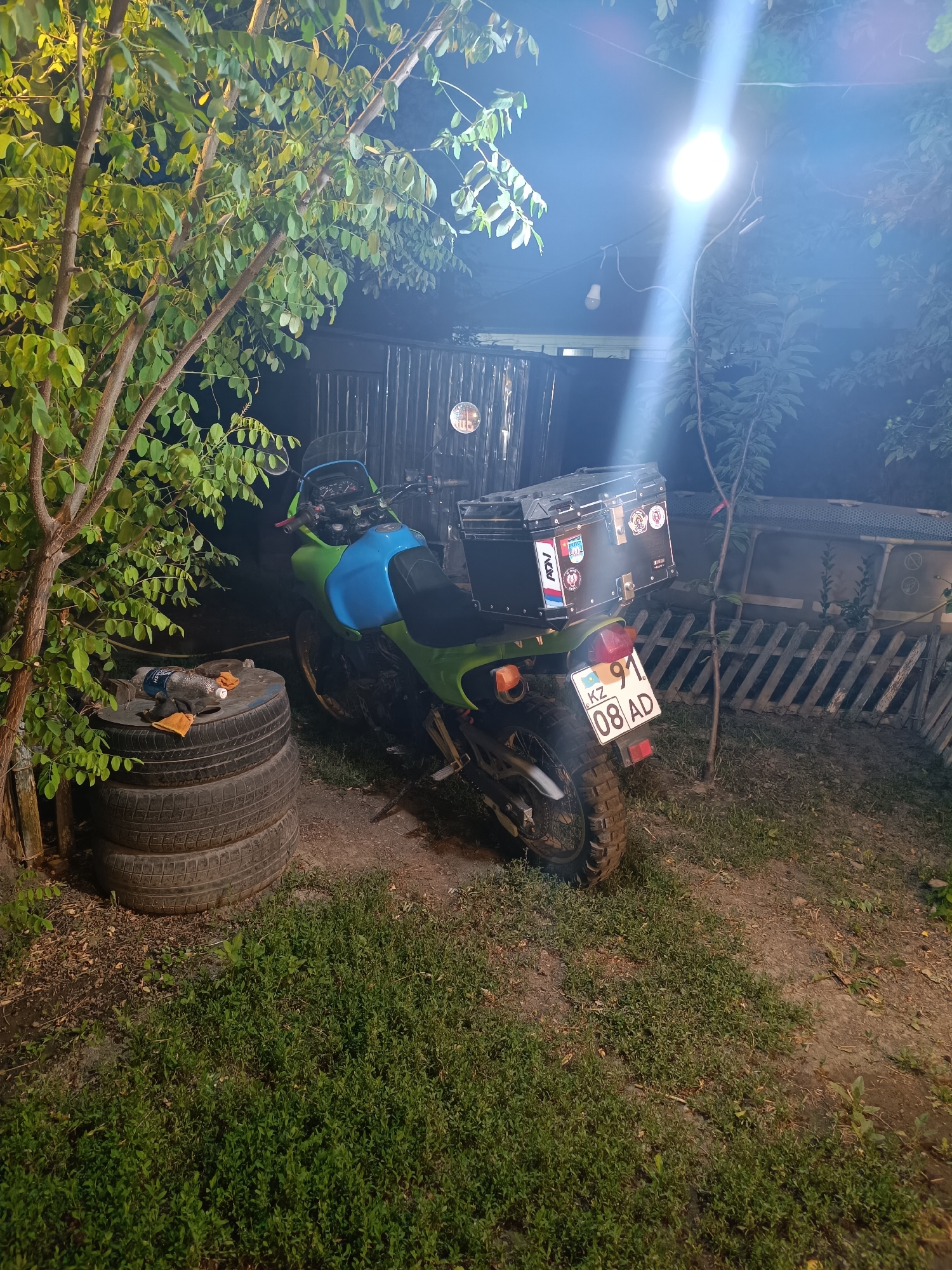 An outdoor scene at night in Taraz, Kazakhstan, features a green and blue motorcycle parked on a dirt and grass surface. The motorcycle is equipped with a black top box adorned with stickers, a windshield, and a license plate displaying "KZ 91 08 AD". To the left of the motorcycle, three stacked tires are visible, with two plastic bottles and two orange cloths placed on top. A tree with green leaves stands to the left, casting shadows. In the background, a corrugated metal fence and a structure are partially illuminated by a bright overhead light. The overall environment suggests a residential or utilitarian outdoor area.