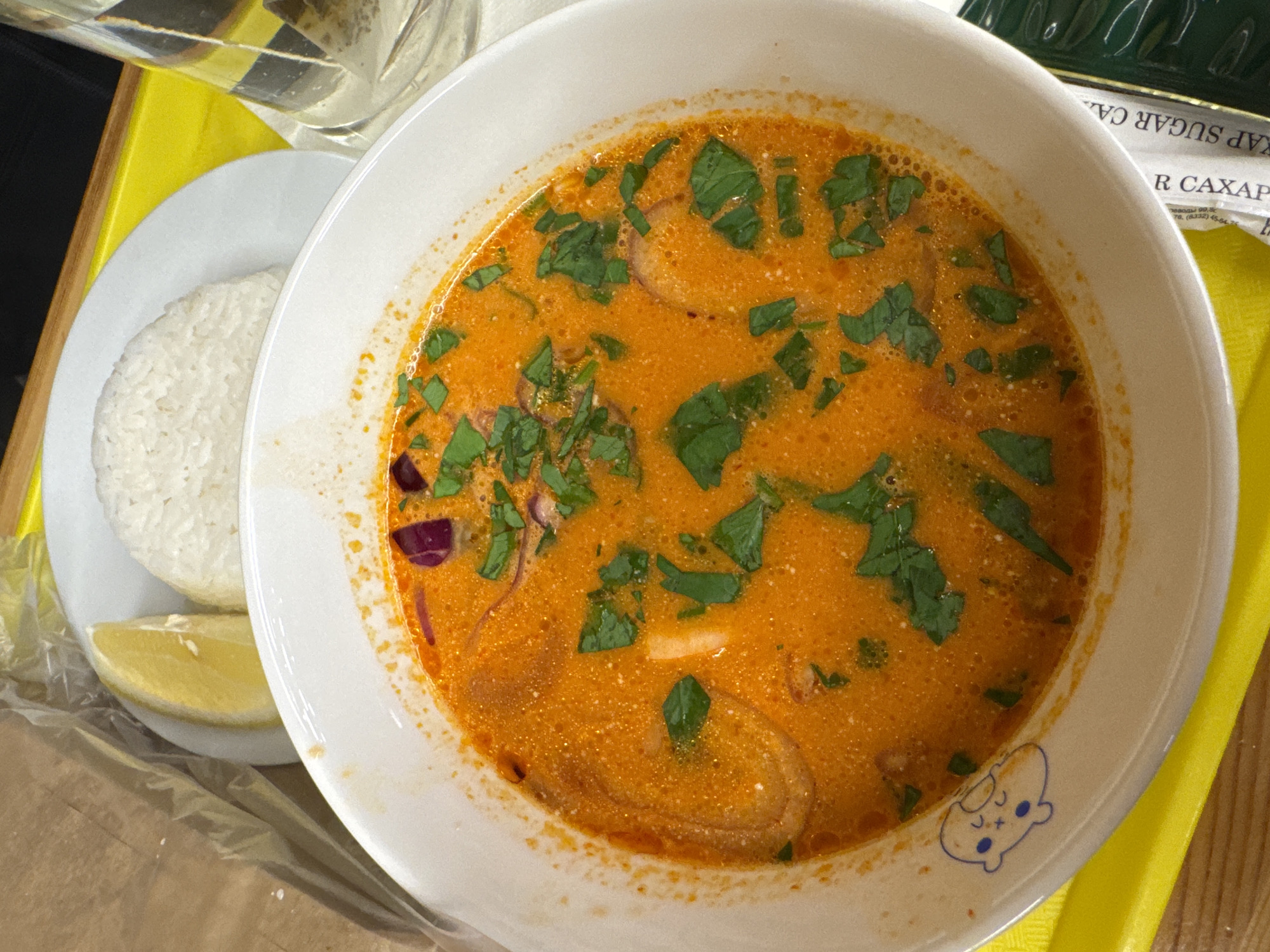 The image shows a bowl of orange-colored soup with green herbs, likely cilantro or basil, scattered on top. Sliced onions and red onion pieces are visible within the soup. To the left of the bowl is a small plate with a mound of white rice and a lemon wedge. The scene is set on a yellow tray, possibly in a restaurant or food service setting. A sugar packet and glass of liquid are visible in the background. There is no indication of the location being Izhevsk, Russia.