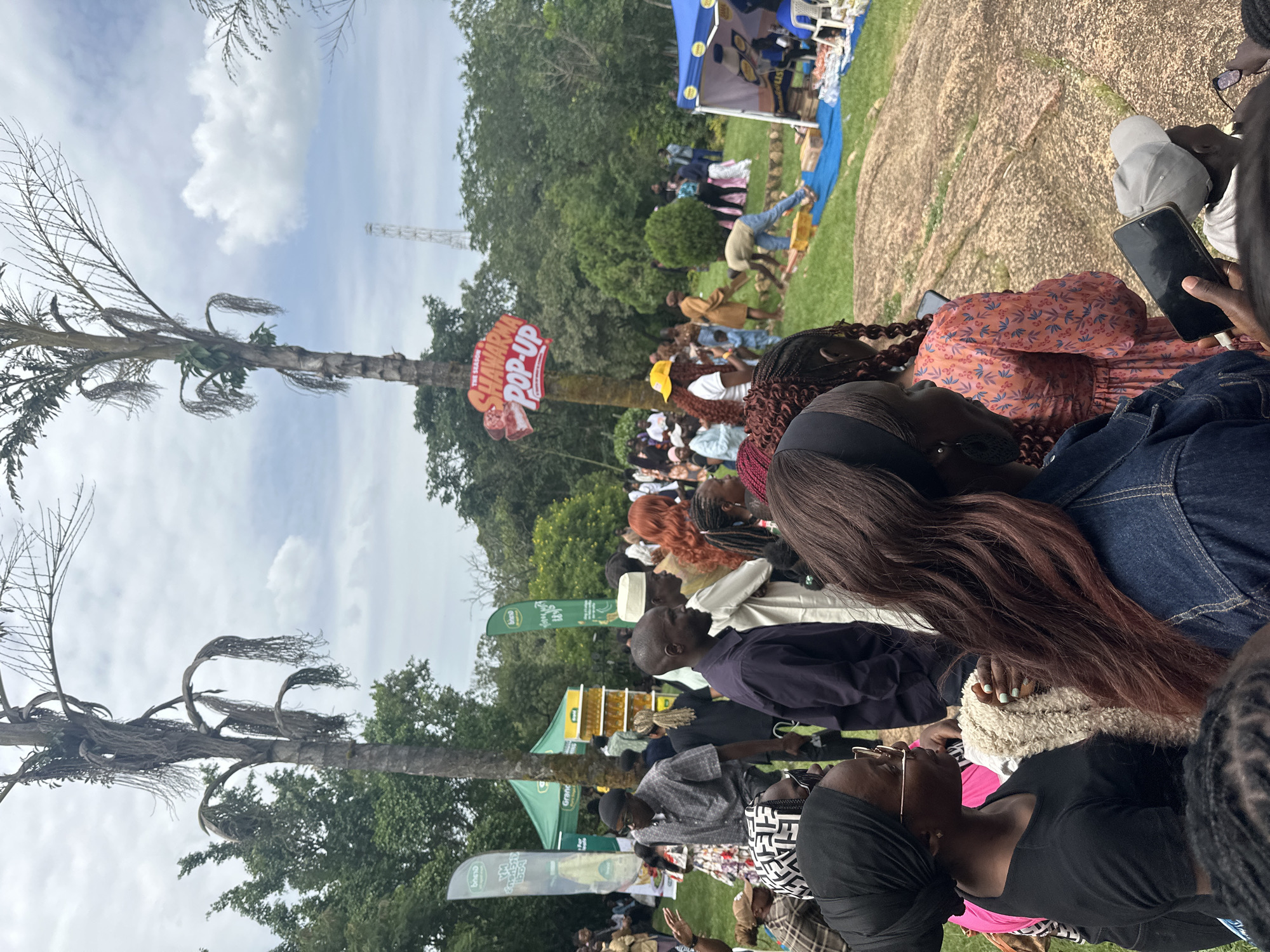 The image captures an outdoor gathering in Jos, Nigeria, characterized by a festive atmosphere. A crowd of people, diverse in attire and ethnicity, are milling around a park-like setting with lush greenery and rocky outcrops. Dominating the background are tall palm trees, one of which has a "Shawarma Pop-Up" sign attached to it. Tents and stalls are visible, suggesting a market or event. People are dressed in a mix of casual and traditional clothing, with some wearing headscarves and traditional caps. The sky is partly cloudy, indicating a temperate day. The overall scene suggests a community event or a local market day in Jos.