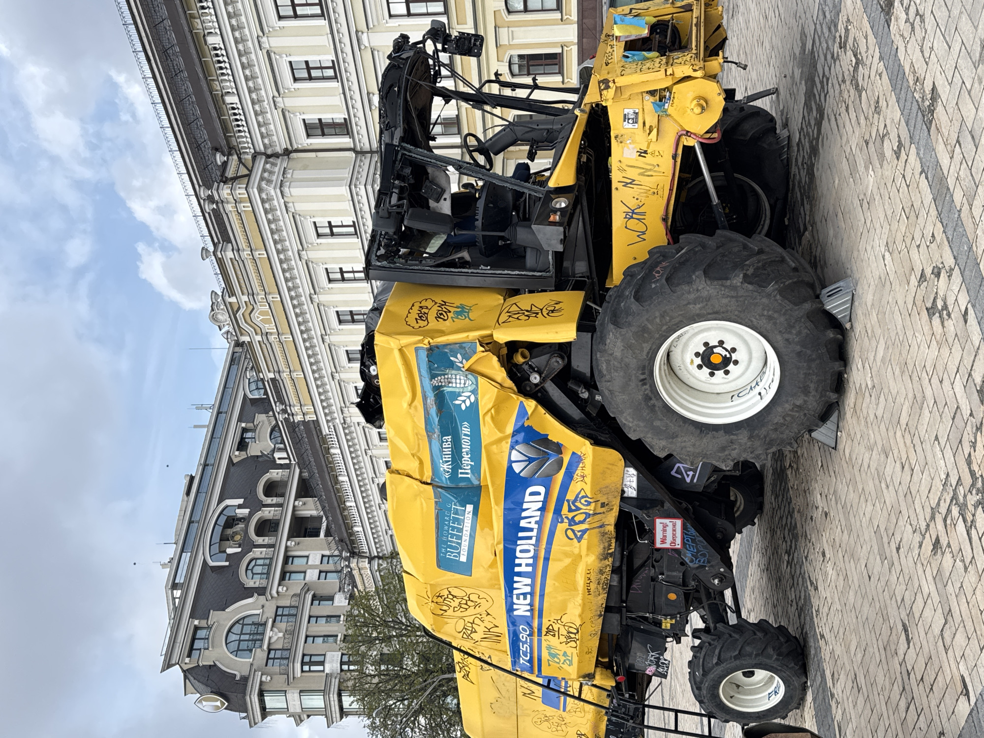 A yellow New Holland combine harvester stands on a paved surface in what appears to be a city square in Kyiv, Ukraine. The harvester is heavily damaged, with graffiti markings covering its exterior. Decals from "The Howard G. Buffett Foundation" and the phrase "Жнива Перемоги" (Harvest of Victory) are visible on the vehicle. The front cabin area is missing or severely damaged, exposing the interior. The tires are large and appear used. Buildings with architectural details are visible in the background. A warning sign, "Warning! Обережно! СМЕРТ" (Warning! Caution! Death), is affixed on the machine.
