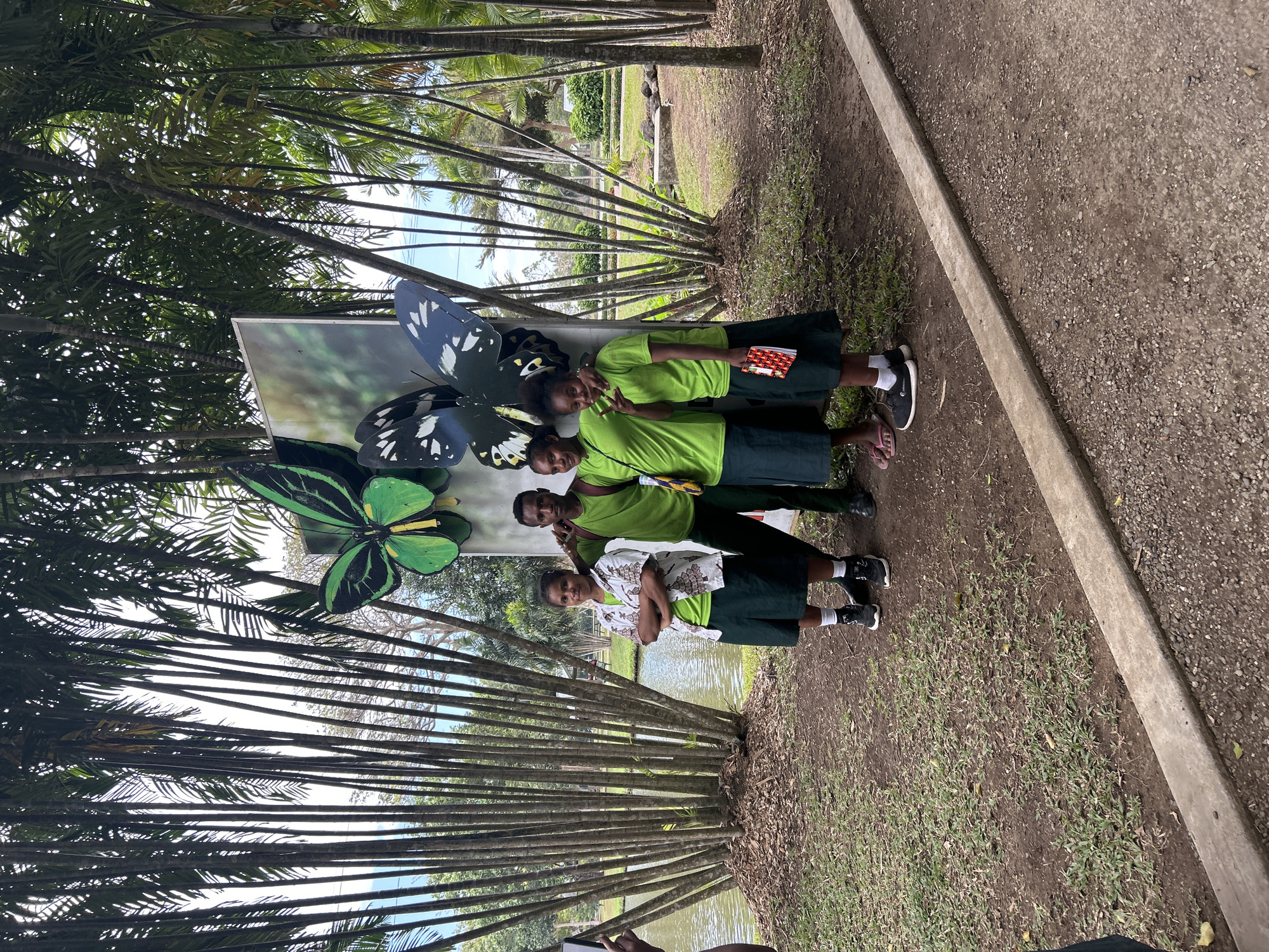 In Tanubada, Papua New Guinea, four individuals, two males and two females, stand in front of a large display featuring two oversized butterflies. The background consists of a calm body of water, dense vegetation with tall, thin trees resembling bamboo, and a paved pathway. The two males, dressed in matching green t-shirts, are positioned in the center of the group. One male is slightly behind the other, with his arm casually resting on the other's shoulder. The female on the left wears a white shirt with a patterned design over a green t-shirt and a dark green skirt. She has her arms crossed. The female on the right is also wearing a green t-shirt and a dark green skirt, and she is holding a small book. The overall scene suggests a tourist or educational outing in a tropical environment.