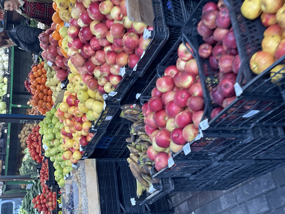 This image depicts a fruit and vegetable stall within a market in Batumi, Georgia. The scene is dominated by a diverse array of produce displayed in black plastic crates.
In the foreground and midground, numerous crates are filled with various apples: primarily red, yellow, and green varieties. Directly to the left of the apples, a stack of black crates contains ripe bananas. Further left, a wooden crate holds light-colored grapes, with another box of darker grapes partially visible.
In the background, additional produce is arranged on shelving units. Large quantities of red tomatoes and green cucumbers are visible to the left, while mounds of orange citrus fruits are present to the right. Beyond these, potatoes, cabbage, and other green vegetables can be seen.
Two adult females are located in the upper right quadrant of the image. One woman, seen from the back and side, wears a black puffer jacket and has red-brown hair. To her immediate right, a second woman, wearing a red and black plaid shirt, is partially visible, smiling and looking towards the right side of the frame. Their interaction appears to be a casual exchange.
The market floor is paved with grey interlocking stones or tiles. Green metal support beams and shelving structures are visible in the background, indicative of a covered market environment. White labels are affixed to some of the produce crates.
