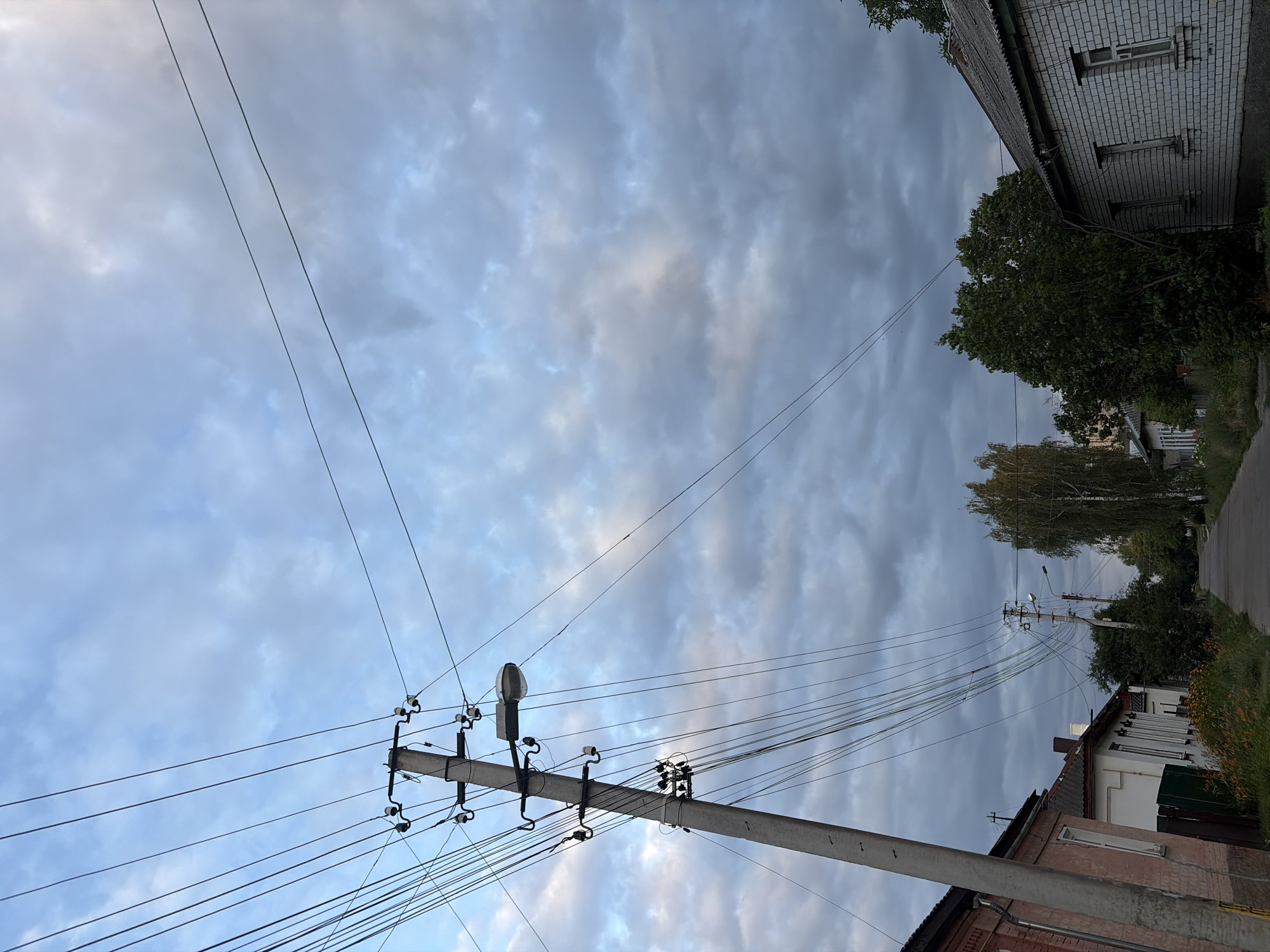 This is a vertical, eye-level shot of a street in Chernihiv, Ukraine, likely in a residential area. The foreground is dominated by a tall, weathered concrete utility pole on the left, supporting numerous overhead electrical wires and a street lamp. The pole is angled slightly to the right. To the left of the pole is the side of a brick building with a light pinkish-red brick facade and white trim around a window. Beyond this building, the roofs of more houses are visible.
The street itself is an asphalt road that recedes into the distance, lined on both sides by houses and trees. On the right side of the street, a white brick house with a grey roof and visible window panes is prominent. Further down the street, more trees and houses can be seen. There are no people visible in the image.
The sky is filled with a mixture of light blue patches and dense, dark grey clouds, suggesting it may be overcast or that a storm is approaching. The overall lighting is muted, indicative of late afternoon or early evening, or a cloudy day. The scene depicts a typical urban streetscape with functional infrastructure and residential architecture.