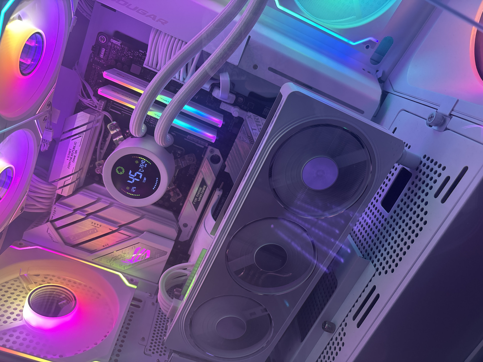 The image shows the interior of a white computer case, illuminated by vibrant purple and pink RGB lighting. A large white graphics card with three fans dominates the lower part of the frame. Above it, a white CPU cooler with a circular digital display is visible, showing a temperature of 45 degrees Celsius. Several sticks of white RAM with RGB lighting are installed on the motherboard, which is partially obscured. White braided cables are neatly managed throughout the case. The surrounding components, including case fans and the motherboard, are also illuminated with colorful RGB effects, creating a visually striking and modern aesthetic. The scene is a close-up interior shot of a custom-built PC.