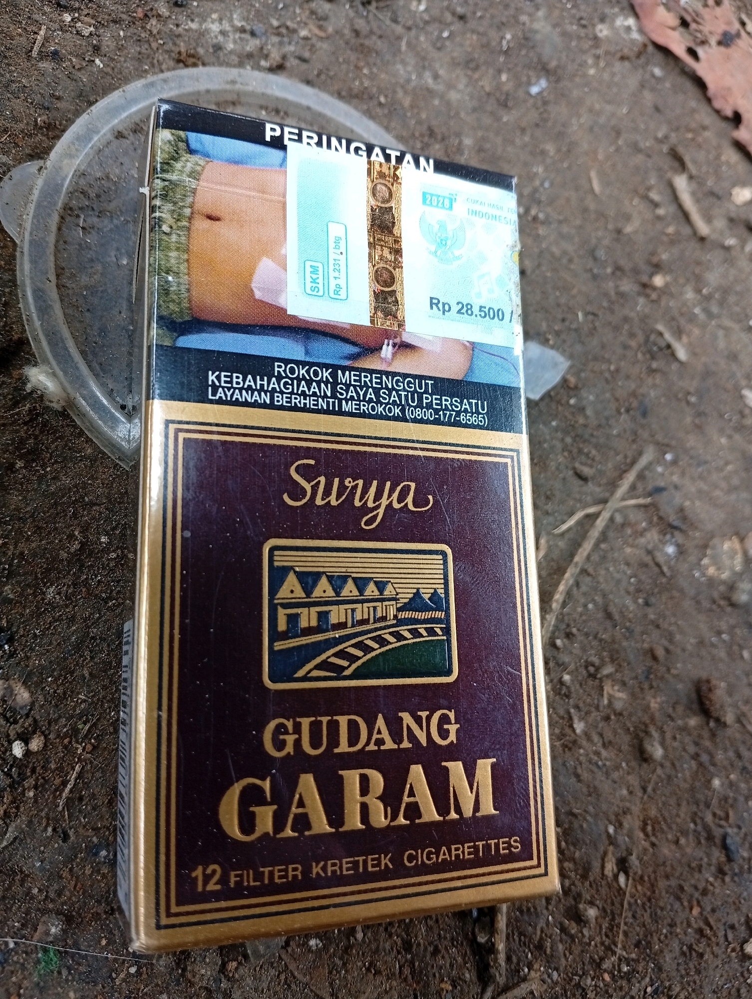 A pack of Surya Gudang Garam 12 filter kretek cigarettes lies on a dirt ground. The pack is predominantly maroon with gold lettering and accents. It features a warning about the health risks of smoking and details about tax stamps. The background consists of dry soil with scattered small pebbles and twigs, suggesting an outdoor, possibly urban or semi-urban environment. There are no people visible in the image. The lighting appears to be natural daylight. The location in Citaman, Indonesia, is not explicitly indicated by any visual cues in the image itself, but is provided as context for the scene.