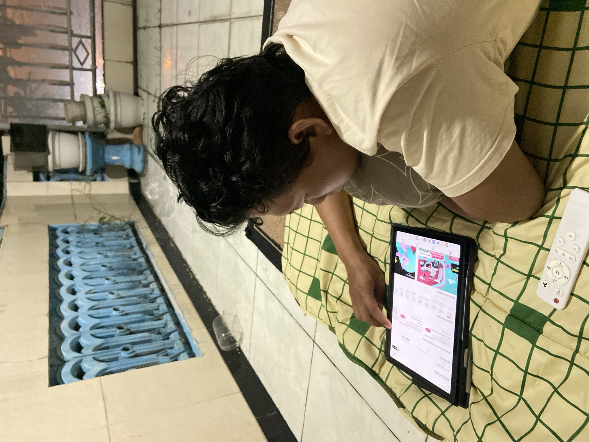 An individual is depicted lying face down on a bed or mat, their head resting on their arms, engrossed in a tablet. They are wearing a light-colored t-shirt and have dark, wavy hair. Their right hand is extended, with a finger touching the tablet screen, indicating active interaction.
The tablet displays the interface of what appears to be the 'VIDIO' streaming application. Visible elements on the screen include various video thumbnails, one featuring a pink background, and categories labeled "Trending" and "Rekomendasi" (Indonesian for Recommendation). To the right of the tablet, a white remote control with buttons marked 'M', 'CC', and 'Q' rests on the bedding. The bedding is light yellow with a green grid pattern.
The setting appears to be an indoor space with a tiled floor. Beyond the person, a blue decorative metal grate is visible at ground level, possibly a ventilation opening or part of a small outdoor area. In the background, planters or pots with greenery are discernable, along with a window featuring a diamond pattern in its frame. A clear plastic cup is on the tiled floor nearby. The lighting suggests it is evening or nighttime. The scene conveys a sense of relaxed media consumption.