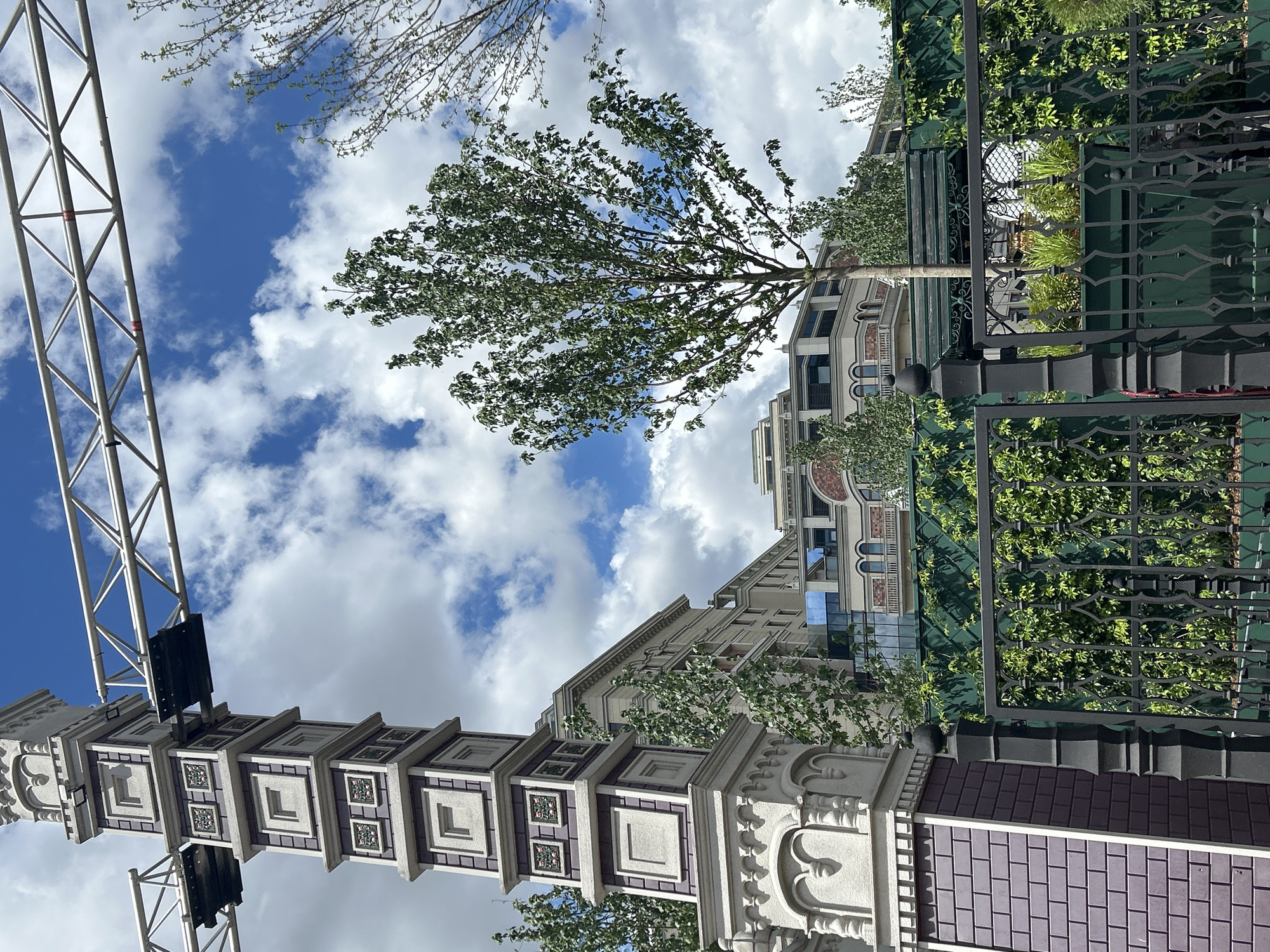 The image shows a street scene in an unknown city, on a sunny day with scattered clouds. The viewpoint is from below, looking upwards.
A tall, ornate structure dominates the left side of the frame. It is constructed of white and purple patterned elements. To its left is a metal truss tower. Lighting fixtures are attached to the metal structure.
In the center of the frame is a multistory building with tan colored walls, arches and rectangular windows. The building is partially obscured by a tree with green leaves.
To the right of the building, a green iron fence is covered with green vines and foliage. Seating can be seen behind the fence.