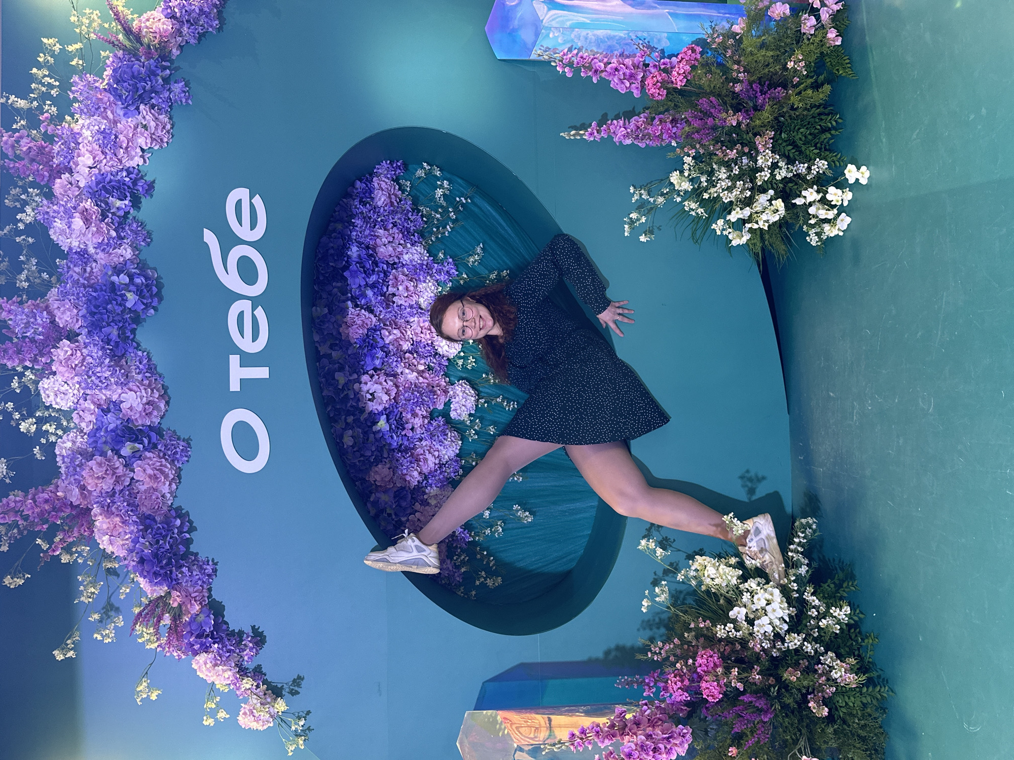 A woman is photographed at an angle that makes it appear she is suspended in the air. She wears a dark dress with light spots and white sneakers, with one leg extended to the left and the other extended to the right. Her arms are outstretched. The background is a blue-green wall decorated with flowers and the Russian text "О тебе". She is positioned within a floral-filled, blue-green oval that is recessed into the wall. The photo was taken in the city of Monchegorsk, Russia.