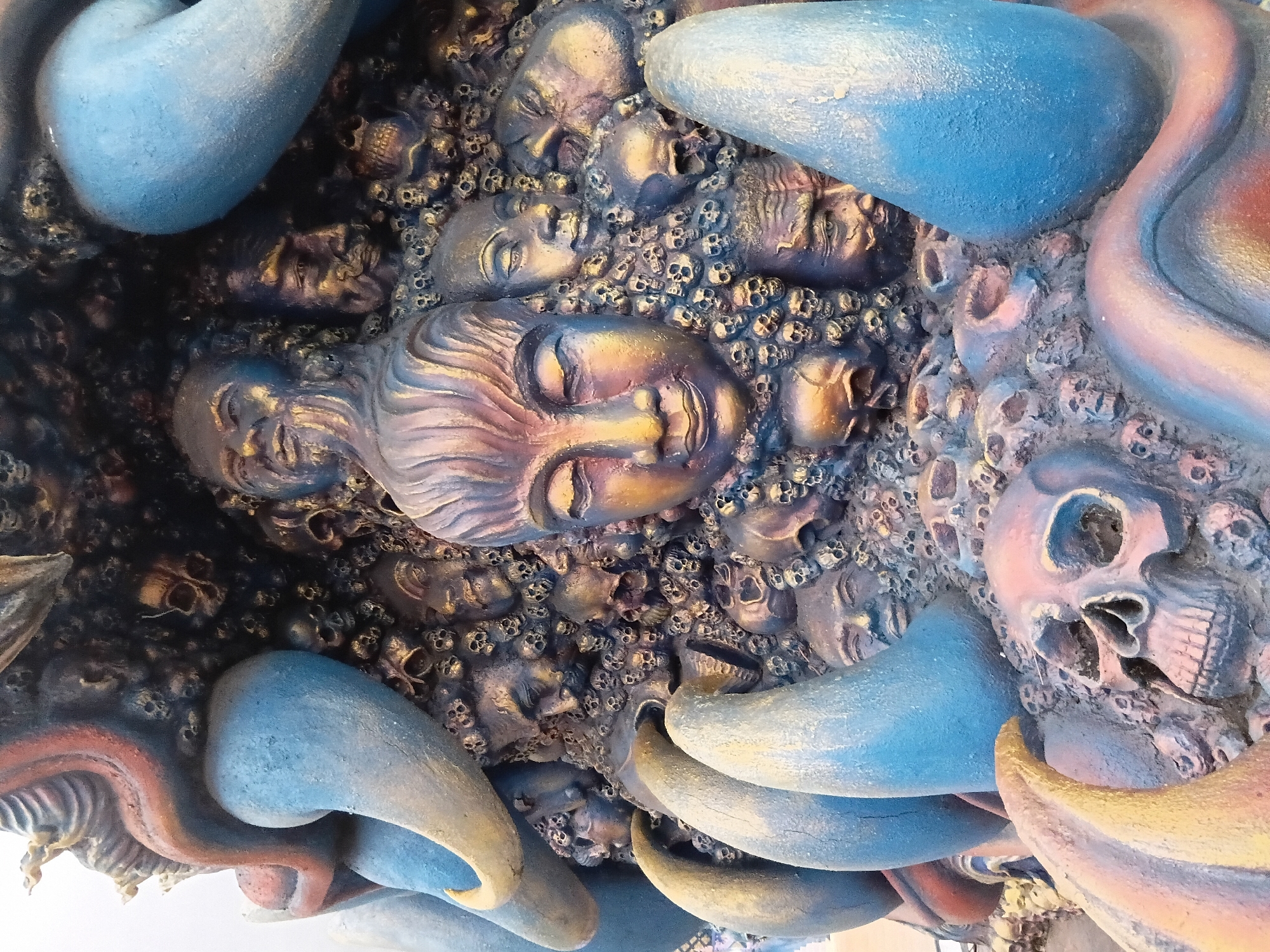 This is a close-up, detailed photograph of a sculpture featuring a large, serene face with closed eyes, surrounded by numerous smaller skulls and distressed human faces. The sculpture is rendered in a metallic, iridescent paint that shifts between gold, blue, and reddish-brown tones, giving it a somewhat otherworldly appearance. Large, curved, blue and gold structures, possibly parts of a larger monstrous form or horns, frame the central composition. The sculpture appears to be a detail from a larger artwork, possibly a shrine or a monument, given the intricate and somewhat macabre subject matter.
This sculpture is located at the "Baan Dam Museum" (Black House Museum) in the city of Chiang Rai, Thailand. The museum itself is an art installation created by the Thai artist Thawan Duchanee, known for his dark, powerful, and often unsettling artworks that explore themes of death, suffering, and the human condition. The Baan Dam Museum is situated approximately 3 kilometers north of Chiang Rai city center.