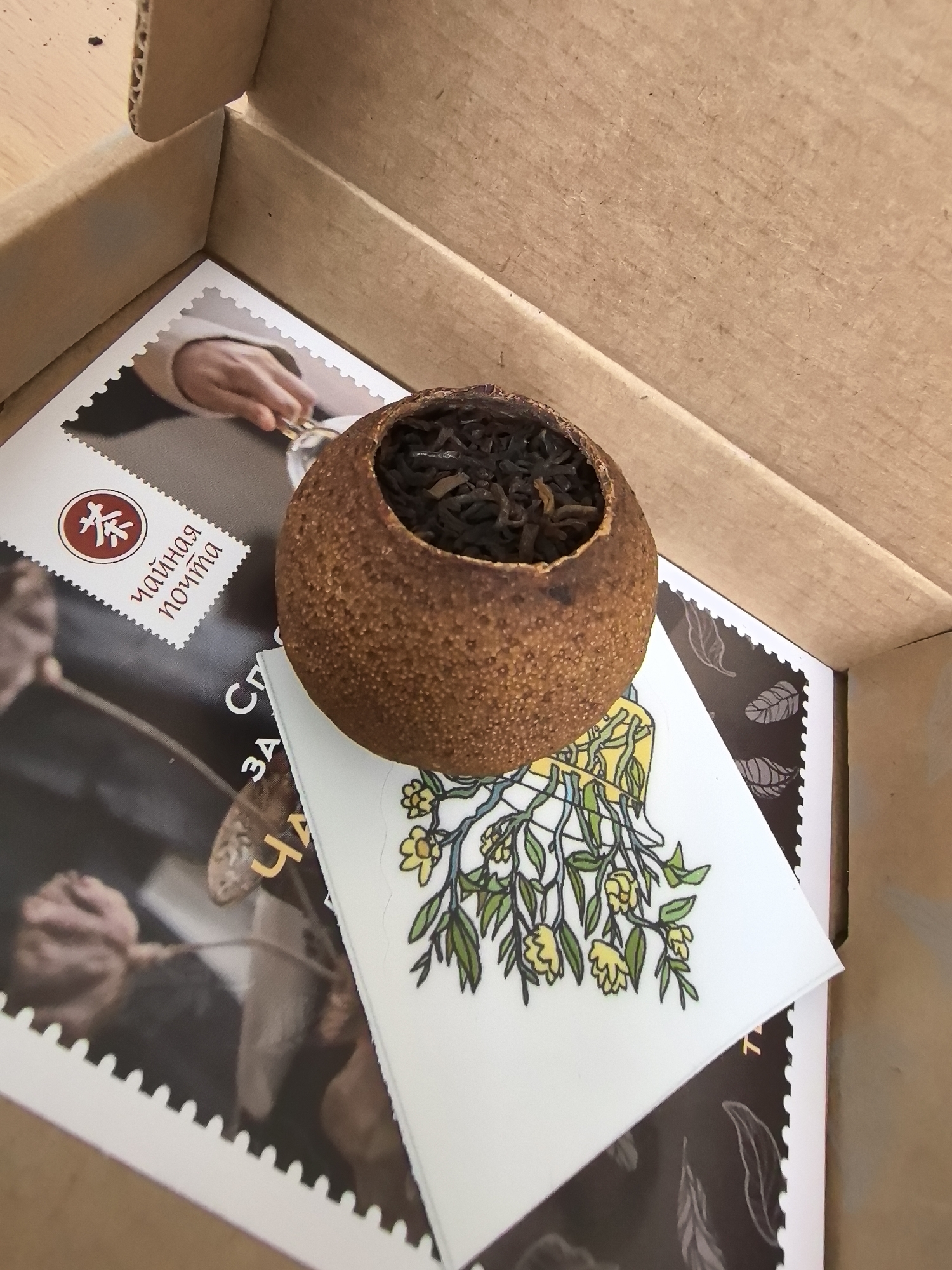 An overhead shot shows a dried tangerine peel filled with loose tea leaves. The tangerine peel is positioned on top of two pieces of paper, which are inside a cardboard box. The top piece of paper is white and features a small illustration of yellow flowers and green leaves. The bottom piece of paper is darker and has an image of a person's hand holding a cup. A small stamp-like image with Cyrillic text "чайная почта" (tea mail) is on the upper left corner of the white paper. The box's interior is visible, revealing a light brown cardboard surface with visible wood grain. The scene appears to be indoors, with the lighting suggesting a natural light source from above. There are no visible people, and the setting is not specific to any city in Belarus.