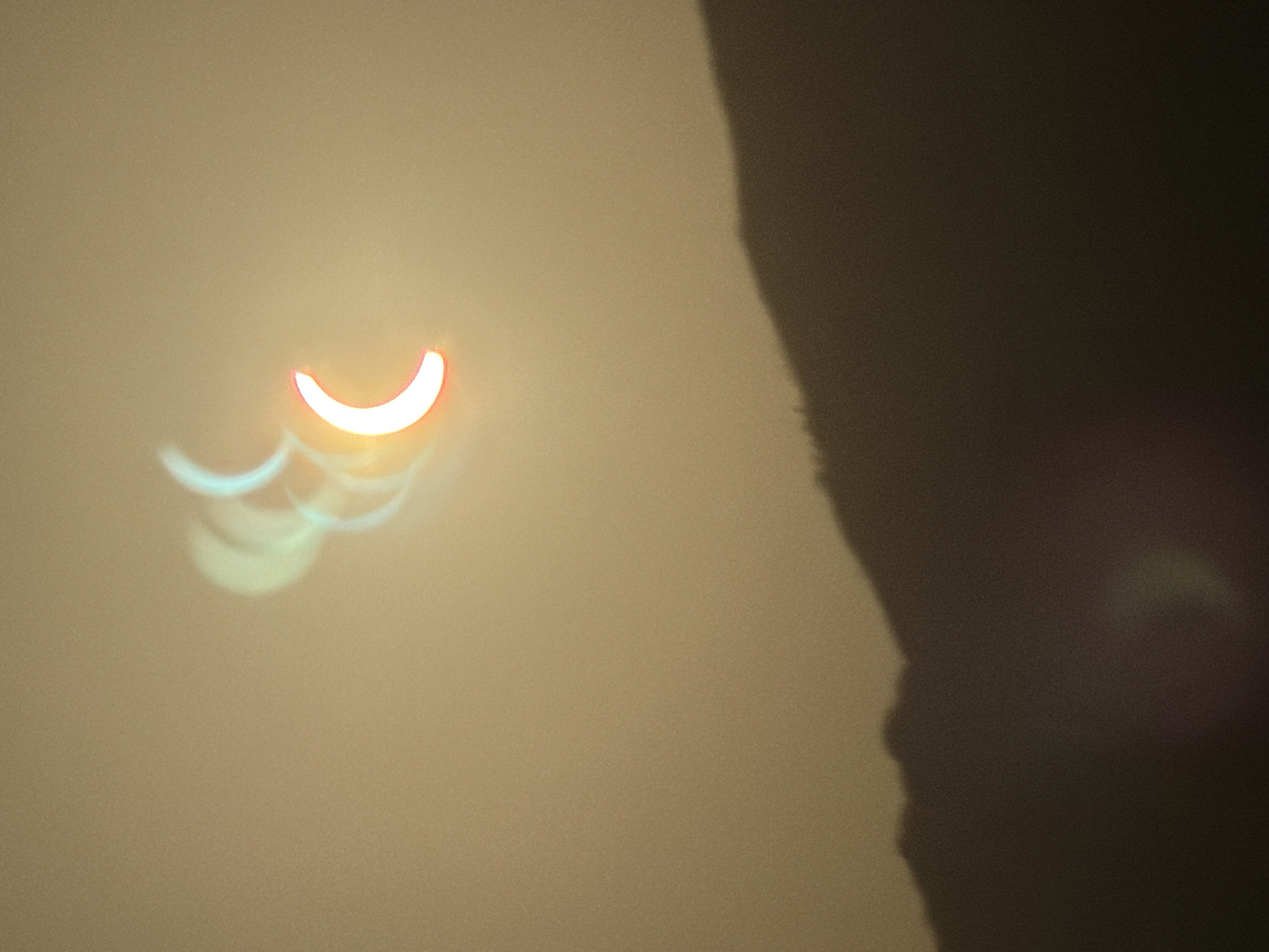 The image shows a partial solar eclipse over a hazy sky with a silhouetted mountain range in the foreground. The sun appears as a crescent shape, with multiple overlapping images suggesting movement or a long exposure. The colors range from bright white and red at the sun's limb to muted yellow and brown in the sky and on the mountains. The overall impression is one of a dramatic celestial event viewed through atmospheric conditions. No specific location in Estepona, Spain, can be identified from the image itself.