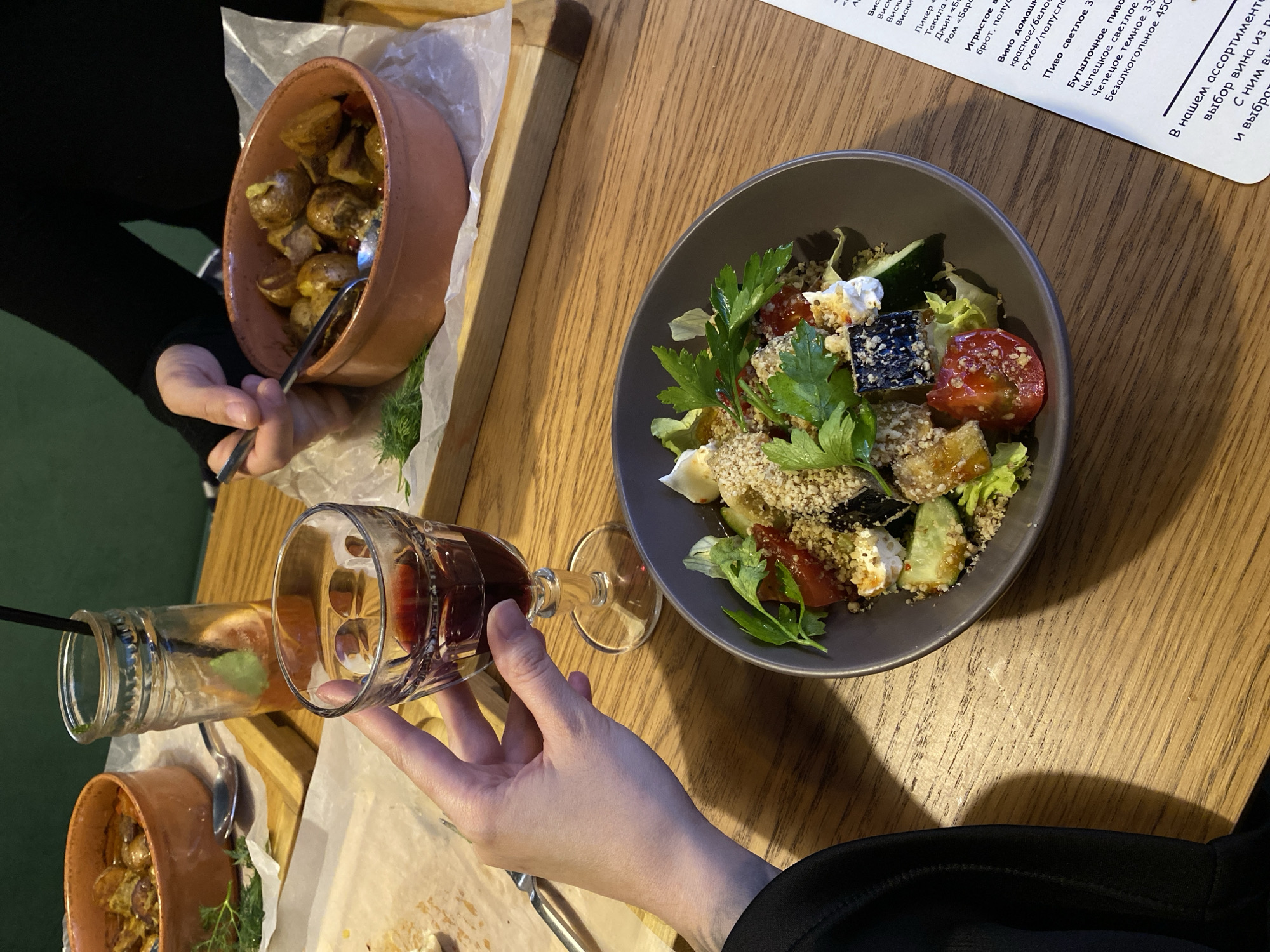Here is a description of the image:
The image depicts a dining table setting, likely in a restaurant, in Votkinsk, Russia, judging by the Cyrillic script in the menu. The table is made of wood and several plates are visible, featuring dishes such as potatoes and a salad with tomatoes, eggplant, and herbs. A glass of red wine is being held by a person's hand. Another person is seen eating from a clay pot with a spoon.
