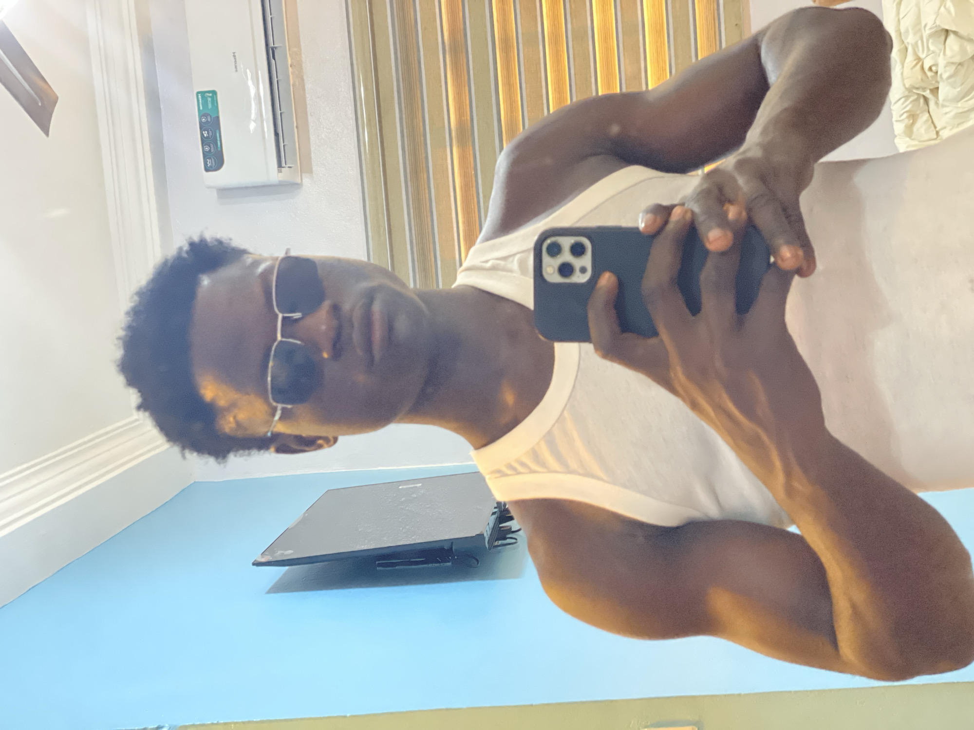 A man with dark skin and short curly hair wearing sunglasses and a white tank top is holding a black smartphone, likely taking a selfie. He is positioned indoors, with a light blue wall, a black flat-screen TV mounted on the wall, and a white air conditioning unit visible. The background features vertical blinds and a glimpse of what appears to be folded white material. The location is indoors, possibly in a room in Badagry, Nigeria.