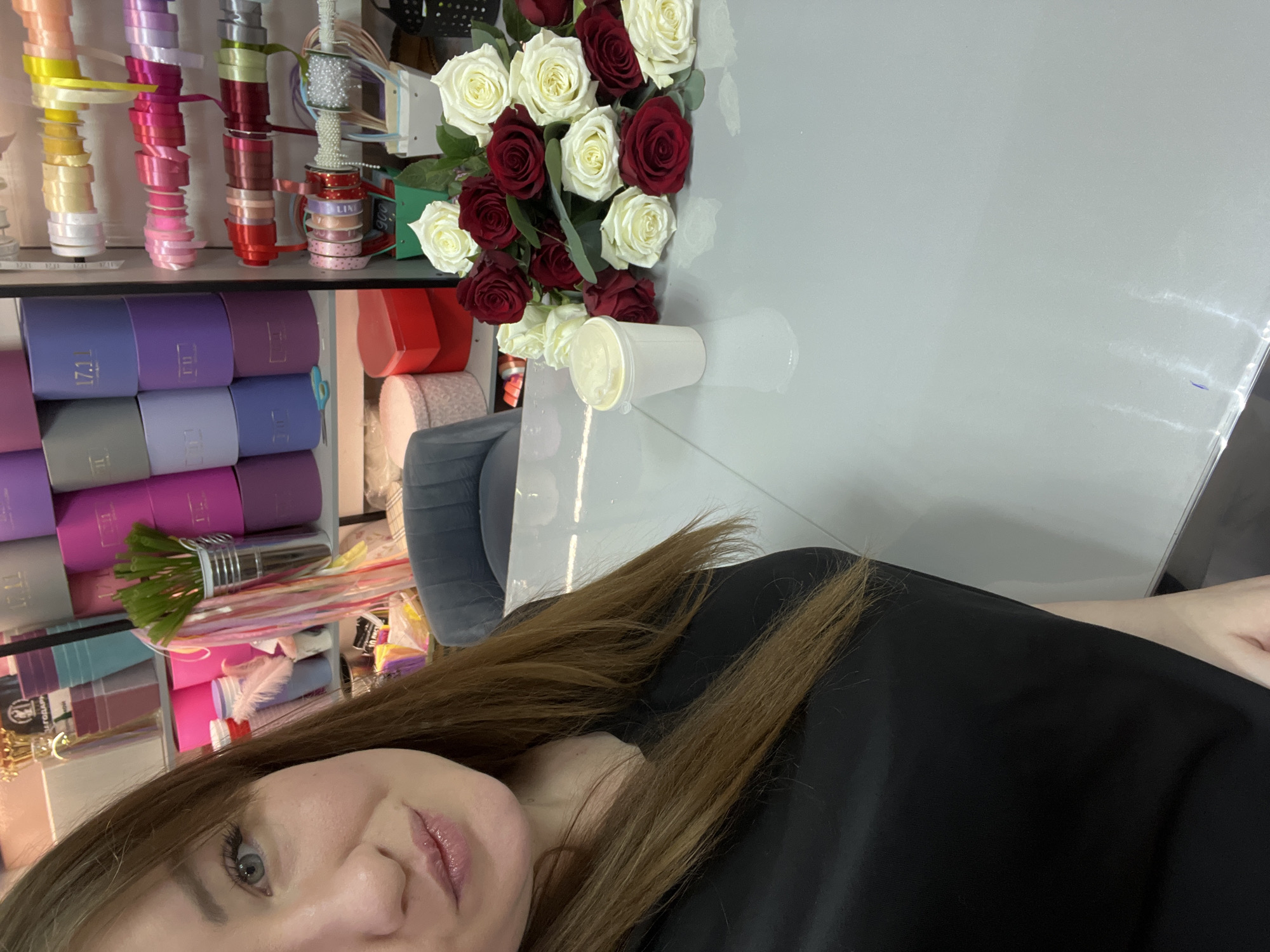A selfie taken in a florist shop in the city of Chaikovsky, Russia. The woman has light skin, brown hair, and is wearing a black shirt. To the left is a display of gift ribbons, boxes and wrapping materials. On a table to the right are a bouquet of red and white roses and a disposable coffee cup.
