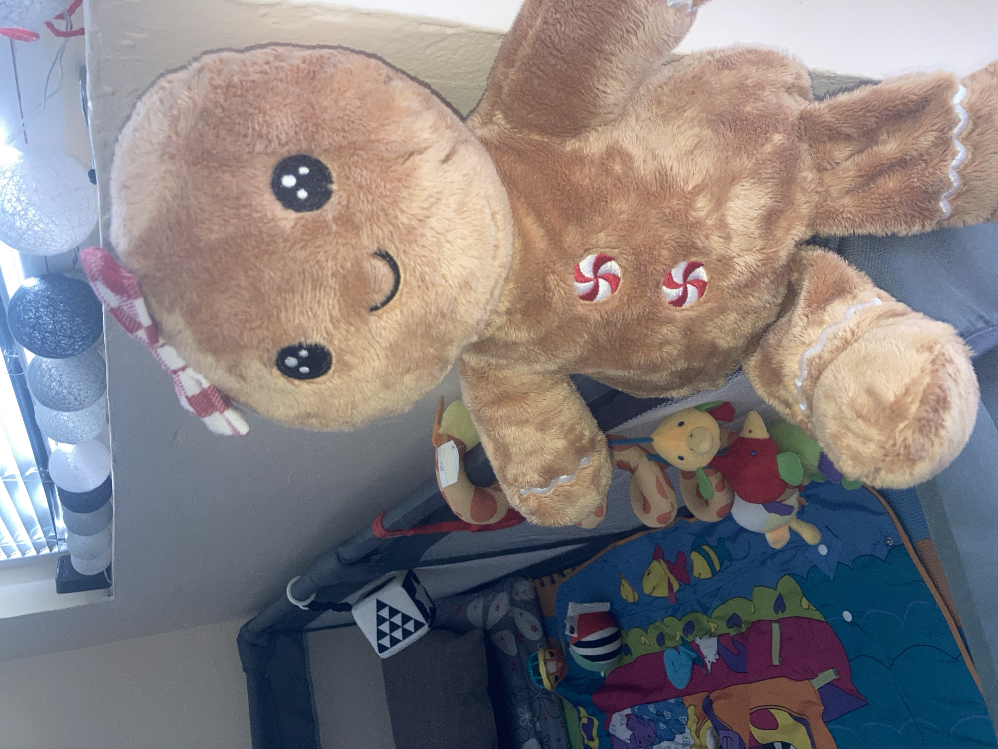 A close-up, eye-level, indoor shot shows a stuffed gingerbread man toy positioned in the foreground. The toy has a light brown fuzzy texture, black button eyes, a stitched smile, a red and white checkered bow on its head, and red and white swirl decorations resembling candy on its chest and arms. To the left of the gingerbread man is a playpen with various baby toys. Among these toys are a black and white cube with triangle patterns, a blue play mat with colorful animal graphics, and several smaller plush toys including a yellow duck, a red bird, and a green and white striped object. In the background, a window with horizontal blinds is visible, and decorative string lights shaped like spheres are arranged along the windowsill. The overall setting appears to be a nursery or playroom. No specific location in Burgas, Bulgaria is discernible from the visual information provided in the image.