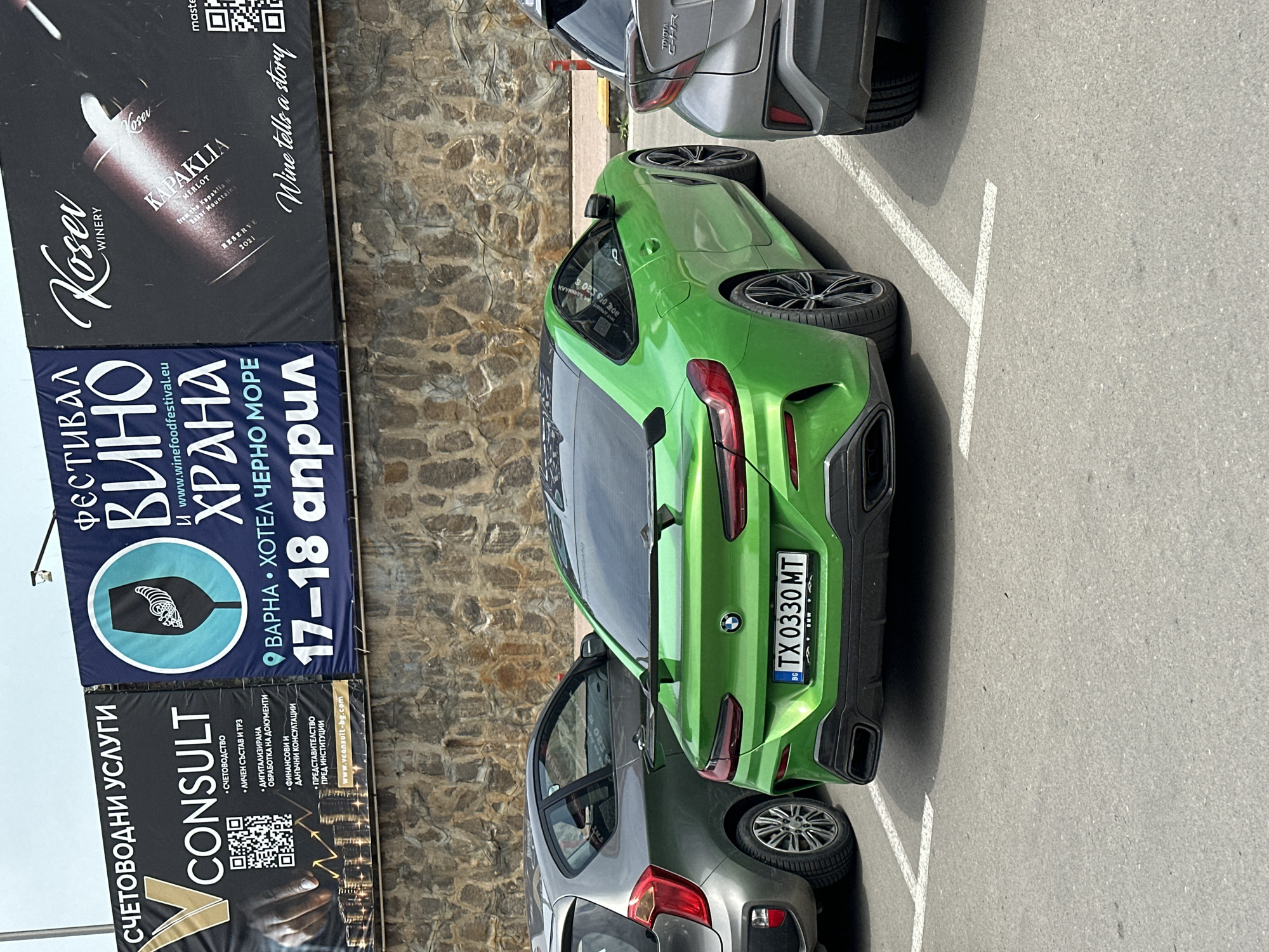 The image shows an outdoor scene in a parking area, likely in Varna, Bulgaria, under bright daylight. A striking bright green sports car, which appears to be a BMW, is parked with its rear facing the viewer. Its Bulgarian license plate reads "TX 0330 MT". A silver Toyota C-HR hatchback is parked partially behind the green car, and another grey hatchback is visible further back.
Behind the vehicles, a stone wall serves as a backdrop, above which three large billboards are mounted. The uppermost billboard advertises "Rusev WINERY" and "KAPAKLIA MERLOT" wine, noting a "2021 RESERVE" and the slogan "Wine tells a story," along with a QR code. Below this, a blue billboard announces a "ФЕСТИВАЛ ВИНО ХРАНА" (Wine Food Festival) at "ВАРНА • ХОТЕЛ ЧЕРНО МОРЕ" (Varna • Hotel Cherno More) on "17-18 април" (17-18 April), with the website `www.winefoodfestival.eu`. The third billboard, on the bottom left, is for "СЧЕТОВОДНИ УСЛУГИ CONSULT" (Accounting Services Consult), listing services such as accounting, HR, digitalization, financial and tax consulting, and representation before institutions, with the website `www.vconsult-bg.com` and a QR code. The presence of Bulgarian text on the billboards and the license plate confirms the location context.