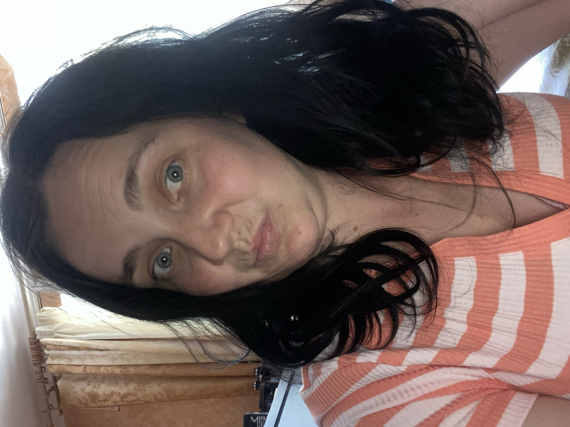 The image is a close-up, low-angle, tilted selfie of a woman with long, dark hair and light blue eyes. She is wearing an orange and white striped shirt. Her face is slightly contorted, with her lips pursed and her eyes looking towards the camera. Her skin appears fair with some freckles visible. The background is blurred, showing what appears to be a curtain or drapes in neutral tones, and a stack of papers or fabric rolls. The lighting suggests it might be daytime, possibly indoors with natural light from a window. The overall emotion conveyed is playful or perhaps a bit mischievous due to her facial expression. There is no visible text in the image.