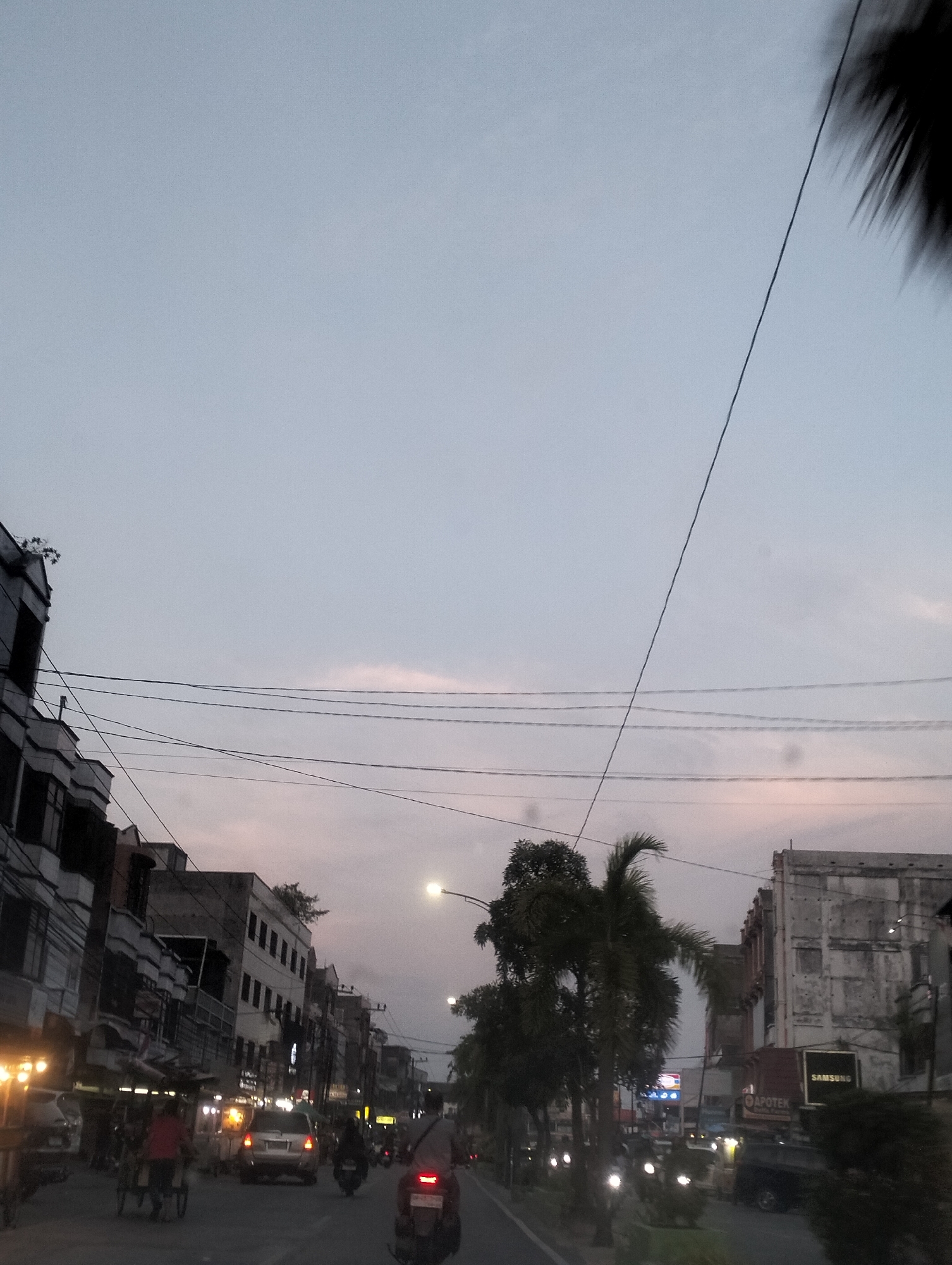 The image depicts a street scene in Dumai City, Indonesia, during twilight hours. The sky is a pale blue with faint hues of pink, indicating either sunrise or sunset. Power lines crisscross the sky above buildings lining both sides of the road. The buildings appear to be a mix of old and modern structures, with some exhibiting faded paint and weathered facades. On the right side of the street, a building with a "SAMSUNG" sign is visible.
The road is populated with various vehicles, including cars, motorcycles, and a rickshaw. Several motorcycles are in motion, with their headlights and taillights illuminated. A silver car is stopped in traffic, and a rickshaw is being pulled by a person. Pedestrians are also present, though some are indistinct due to the low light conditions. Lights from street lamps and shopfronts illuminate the scene. Trees, including palm trees, are interspersed along the roadside, adding to the tropical atmosphere. The overall impression is one of a moderately busy street in an urban setting during the transition from day to night.