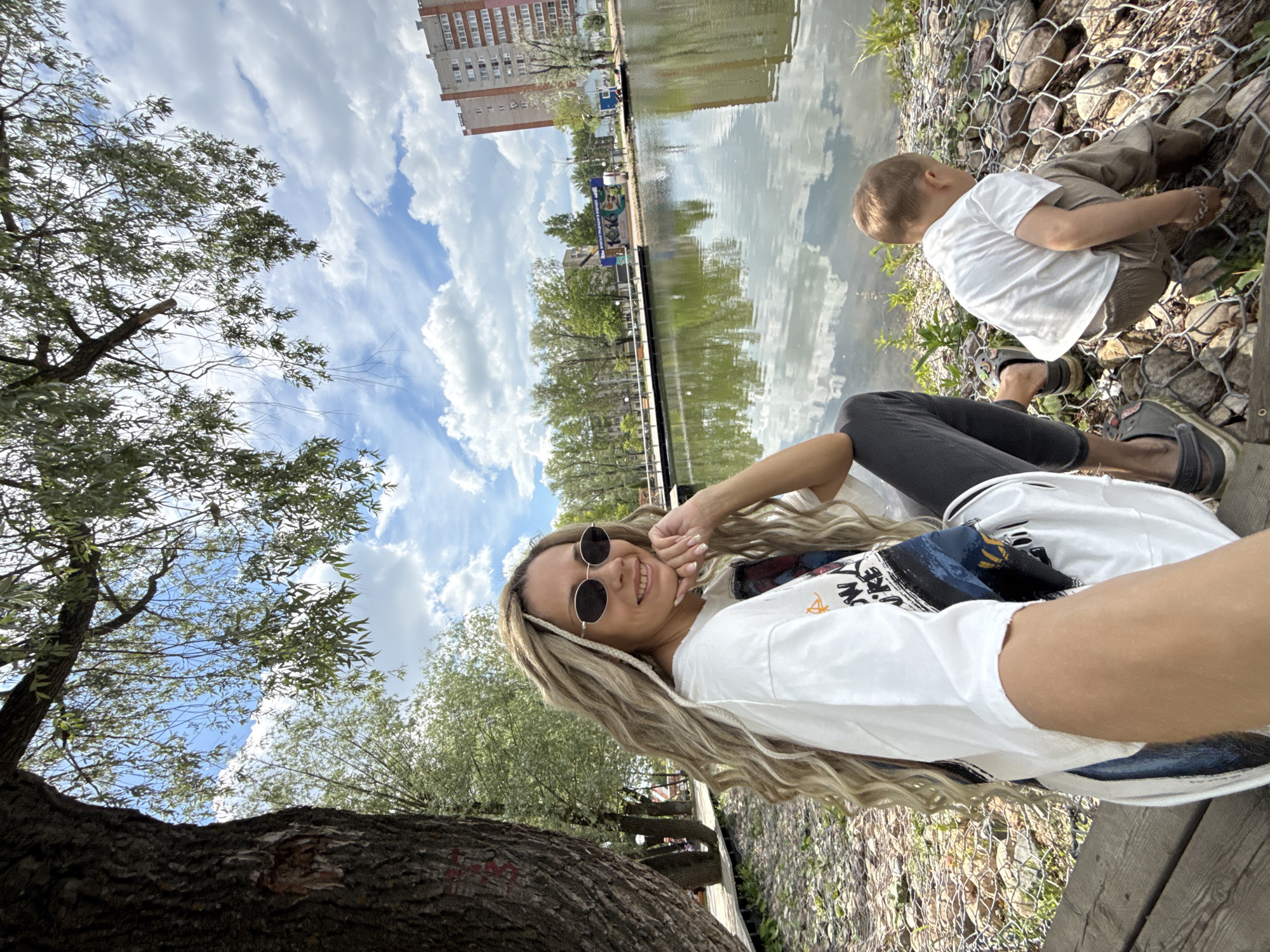 This outdoor image, rotated 90 degrees counter-clockwise, captures a relaxed moment by a body of water in Rusanovo, Russia. In the foreground, a woman with long, wavy blonde hair and dark round sunglasses is taking a selfie, smiling warmly at the camera. She wears a white t-shirt with a graphic design and dark leggings, with a dark bag slung over her shoulder. To her right, a large tree trunk dominates the left side of the frame, with its green leaves visible against the sky.
In the middle ground, a young child, dressed in a white t-shirt, light brown pants, and dark sandals, crouches near the water's edge, seemingly engaged in an activity by the rocks of a gabion wall. The calm water, likely a canal, reflects the cloudy blue sky and distant buildings and trees. Across the water, apartment buildings with red-brick upper stories are visible, along with a structure featuring a colorful mural.
The sky is bright with fluffy white cumulus clouds, suggesting pleasant daytime weather. The overall scene conveys a casual and enjoyable outing, with both individuals focused on their immediate surroundings or interactions. No clearly legible text is visible on clothing or structures.