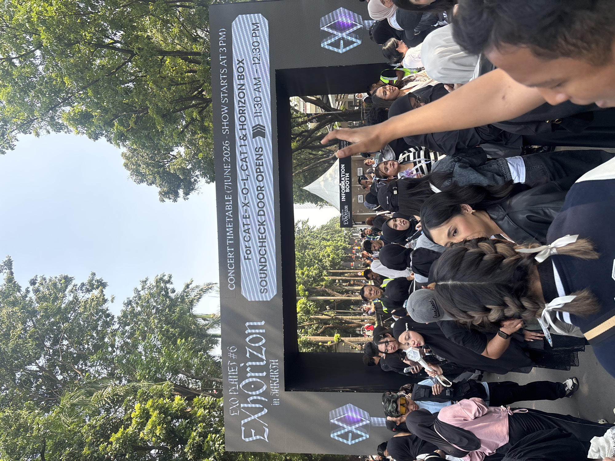 This image captures a dense outdoor gathering of people, primarily young adults, attending a concert event identified by a large, illuminated archway. The archway prominently displays "EXO PLANET #6 EXhOrizon in JAKARTA" and a "CONCERT TIMETABLE" indicating the event date as 7 June 2026, with the show commencing at 3 PM. Further details on the sign state "For CAT E-X-O-L, CAT 1 & HORIZON BOX SOUNDCHECK DOOR OPENS 11:30 AM - 12:30 PM."
The crowd is predominantly dressed in black or dark attire, and numerous individuals are holding white, official EXO lightsticks. In the immediate foreground, the back of a person with braided hair and white ribbons is visible on the left, while on the right, a person's raised arm partially obscures the view. Various attendees in the mid-ground are looking forward, to the side, or interacting with others. A woman in a black hijab is seen holding an EXO lightstick near the center. Security or staff personnel in high-visibility vests are discernible within the crowd.
In the background, beyond the archway, a white tent labeled "INFORMATION BOOTH" is present. The entire scene is set outdoors on a paved surface, bordered by numerous tall, green leafy trees under a clear, light blue sky, indicating bright daylight. The location, "JAKARTA," combined with the scale of the event, strongly suggests a major venue like the Gelora Bung Karno (GBK) complex, which is situated in Central Jakarta, Indonesia.